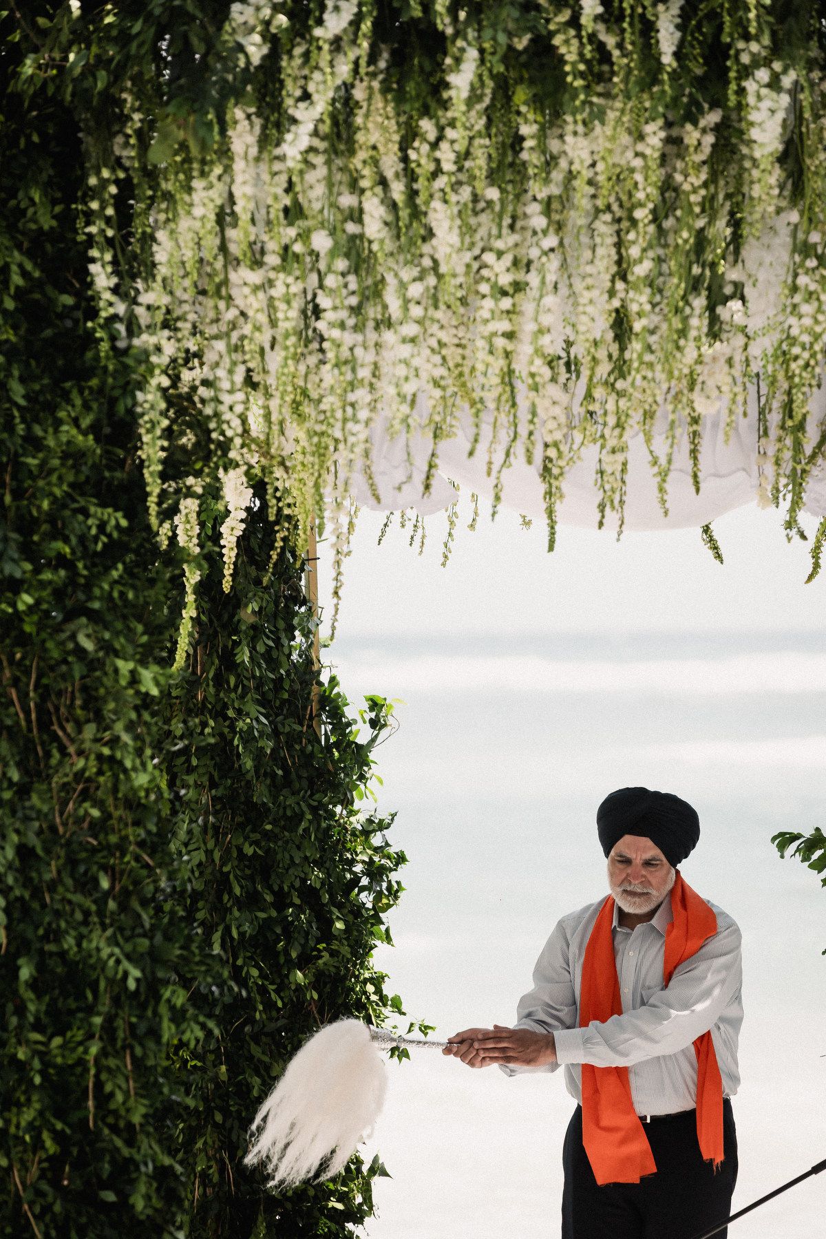 Sikh priest in black turban and orange scarf performs ceremony under floral arch with hanging white wisteria blooms.