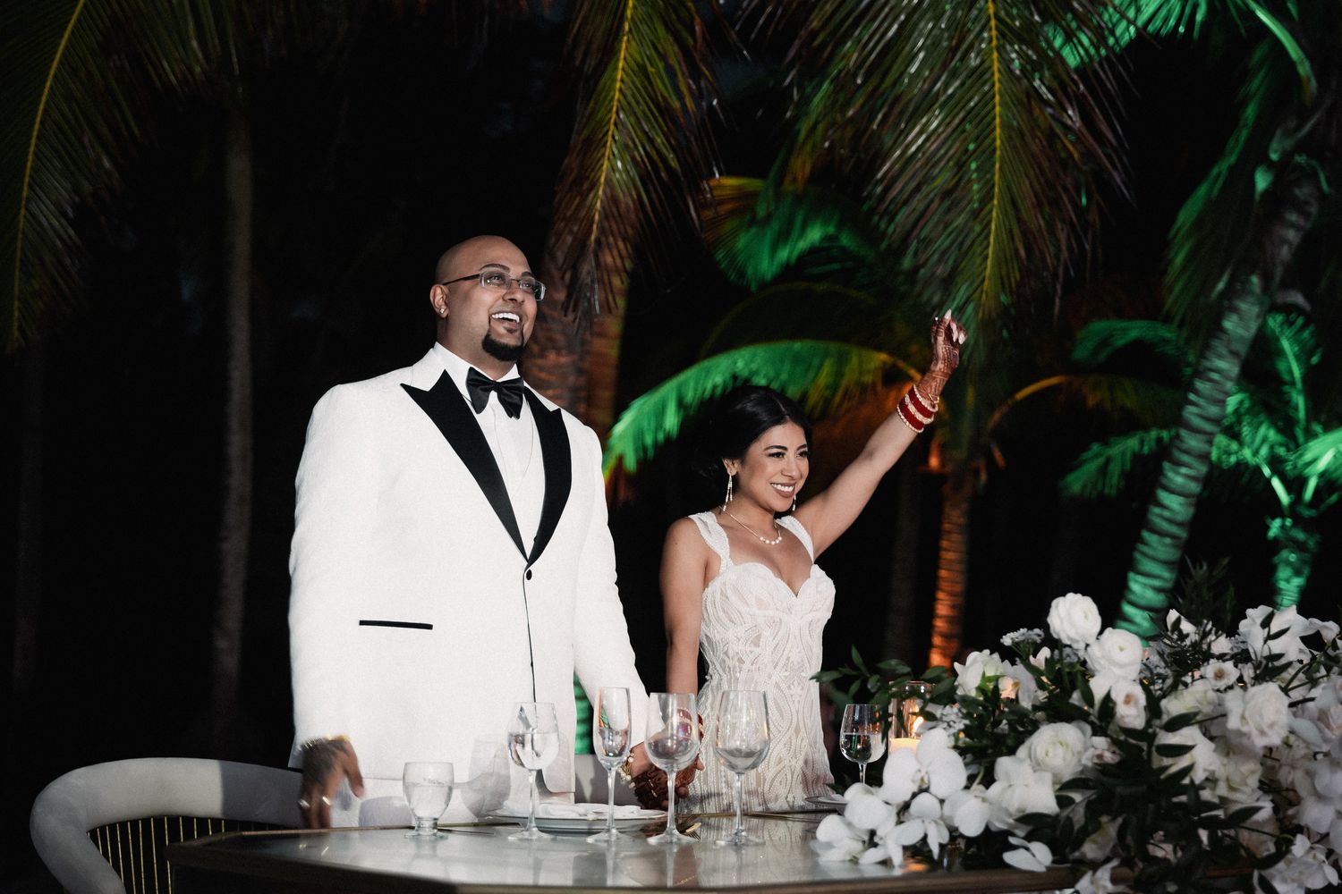 Bride and groom celebrate at sweetheart table during outdoor tropical wedding reception at night.
