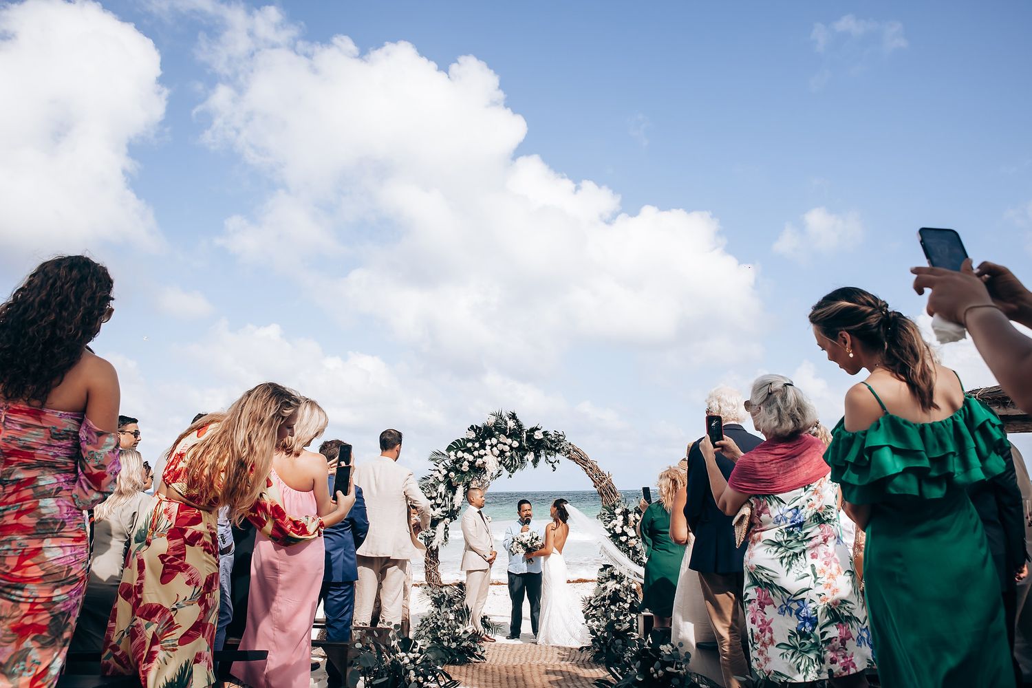 Panoramic view of beach wedding ceremony with guests seated along shoreline.