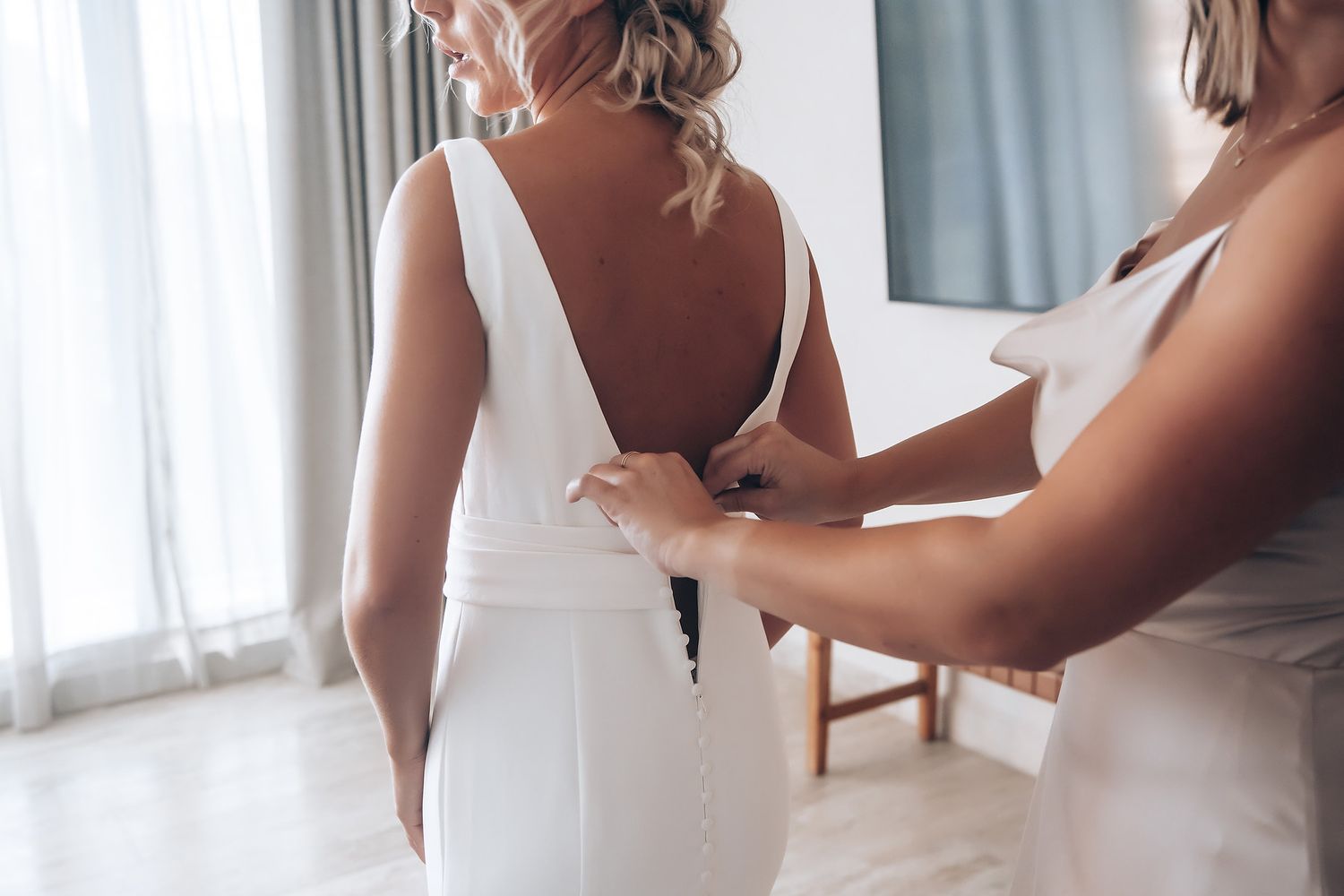 A woman in a white dress gets ready in an elegant dressing room with sheer curtains and natural light.