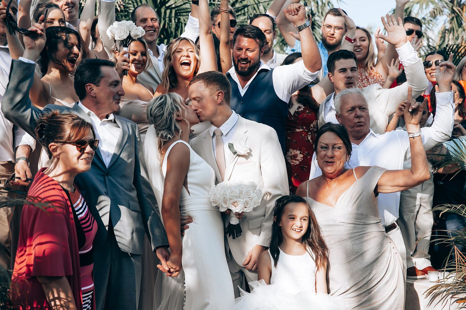 A joyous wedding celebration with guests cheering around the newlyweds outdoors with palm trees in the background.