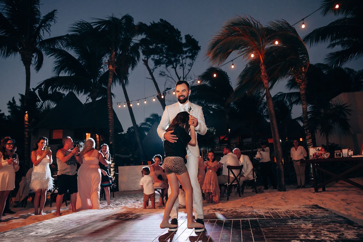 Evening beach wedding reception with string lights and dancing silhouettes against twilight sky.