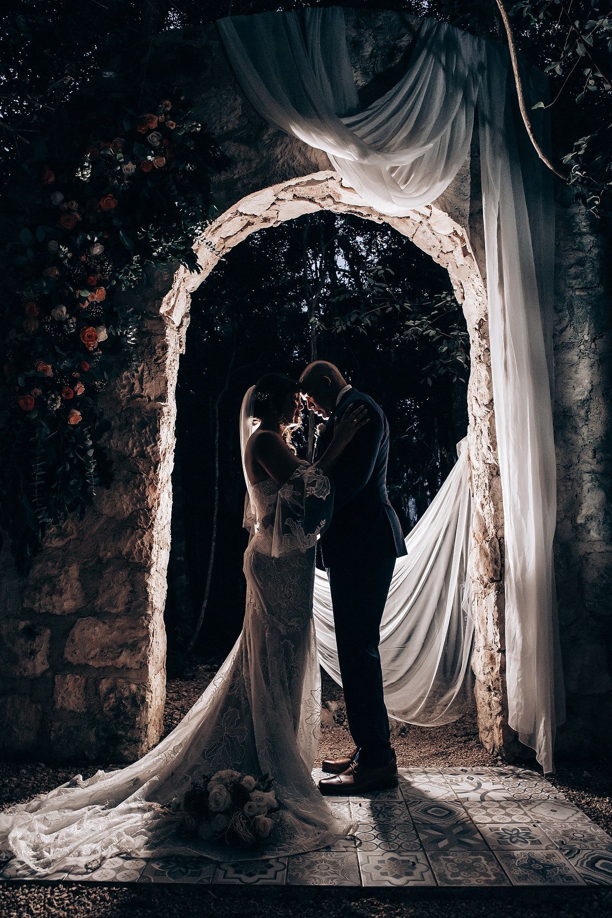 Silhouetted wedding couple poses under illuminated stone archways.