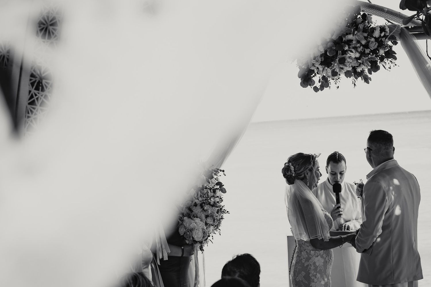 Black and white wedding ceremony shot of bride and groom exchanging vows at floral altar.
