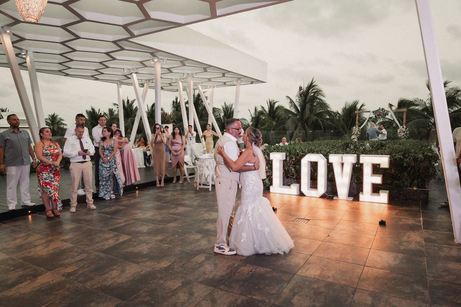 Bride and groom share first dance before illuminated LOVE sign at tropical outdoor wedding reception.