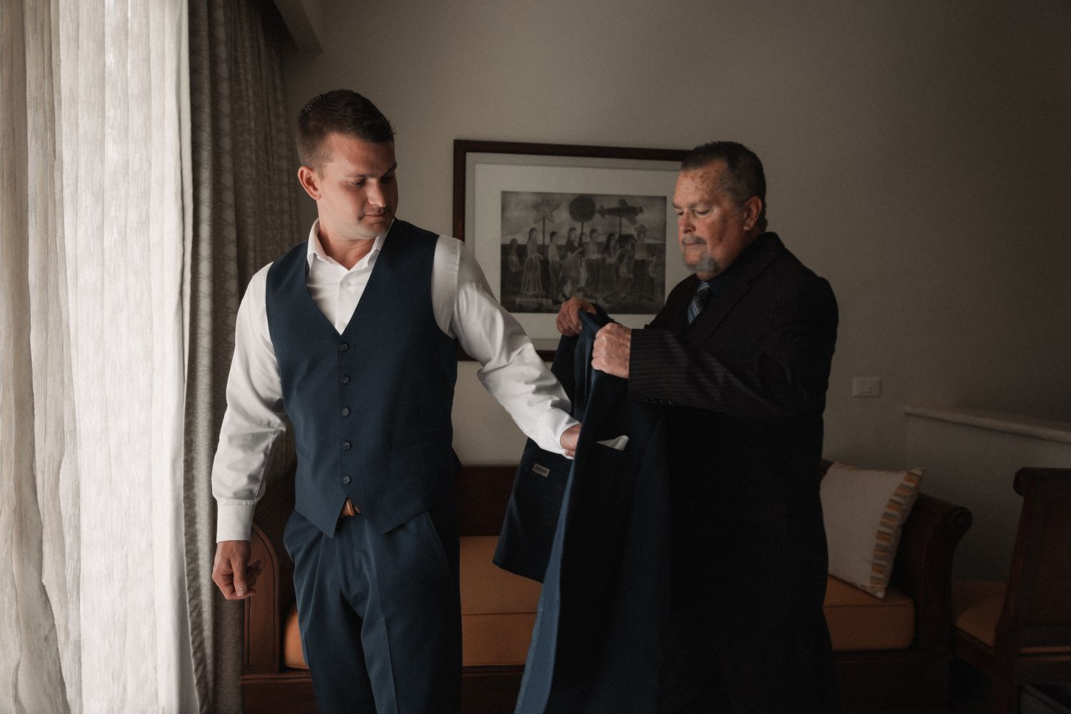 Two men in formal attire, one in a vest and one in black, assist each other getting dressed indoors.