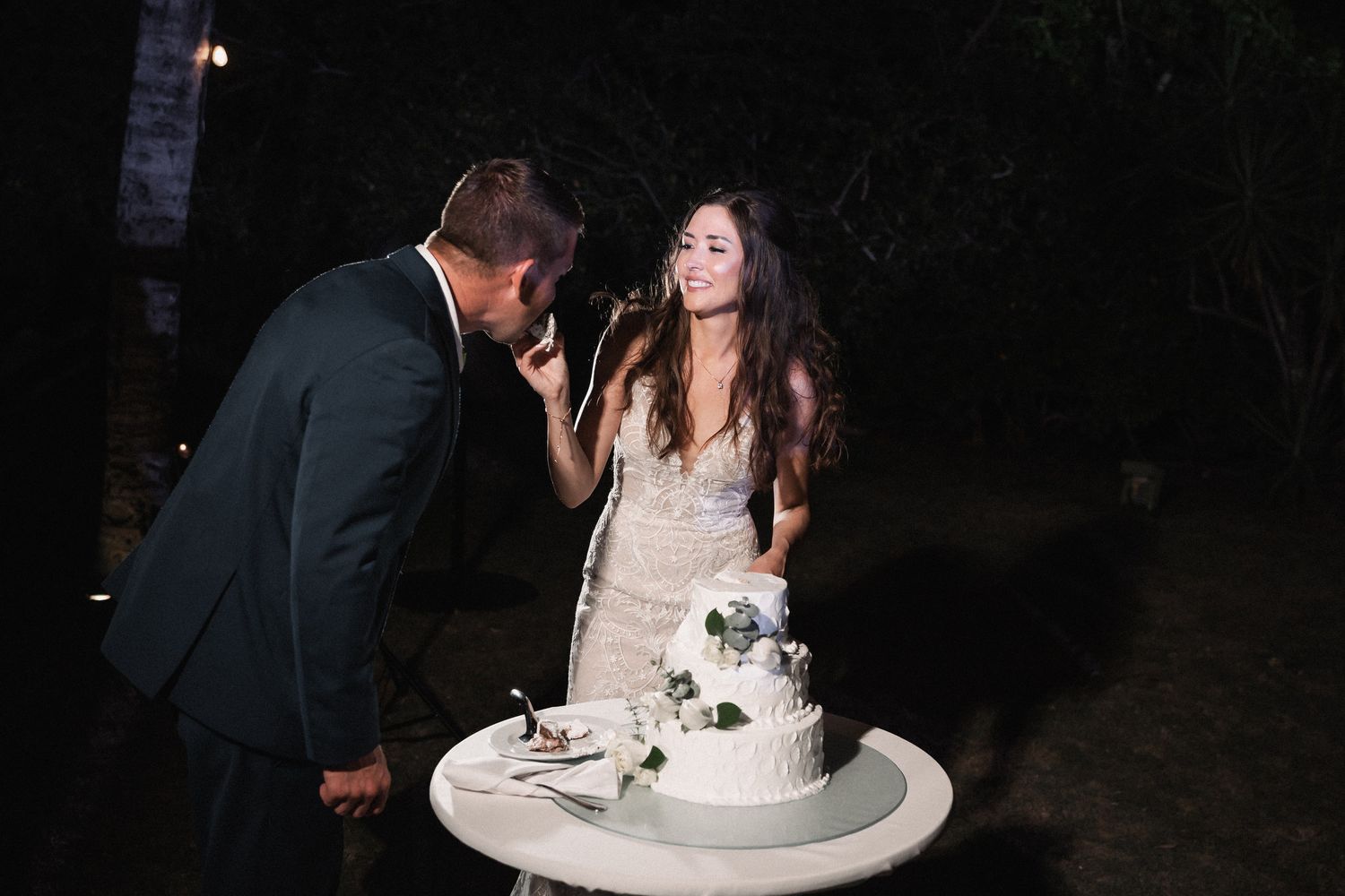 Smiling bride and groom cut their white floral wedding cake together at an outdoor nighttime reception.
