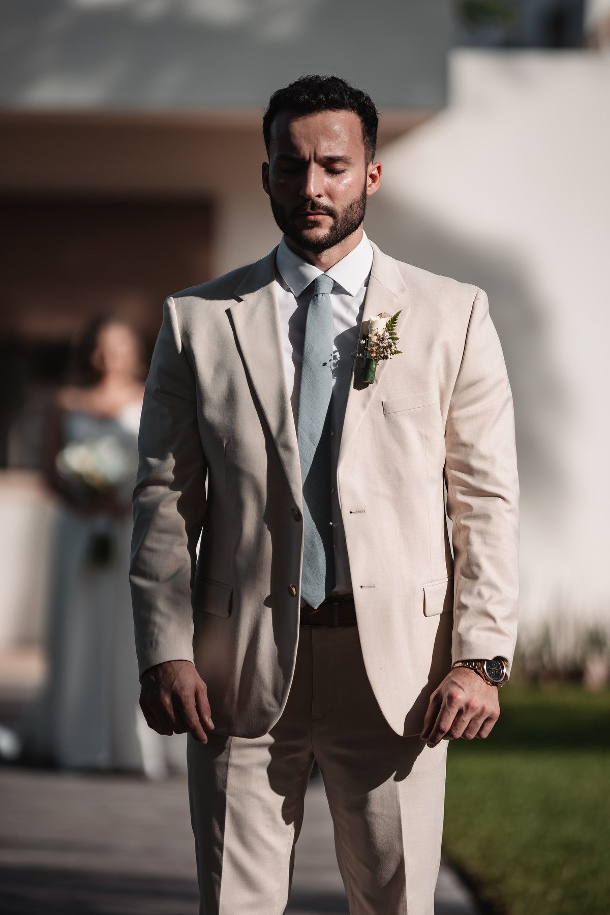 Handsome groom in beige suit with boutonniere standing outdoors in sunlight.