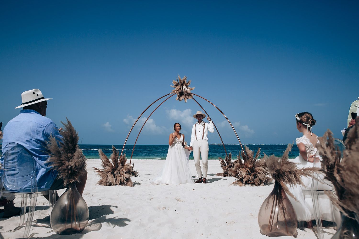 Beach wedding ceremony setup with dried palm decorations against turquoise ocean backdrop.