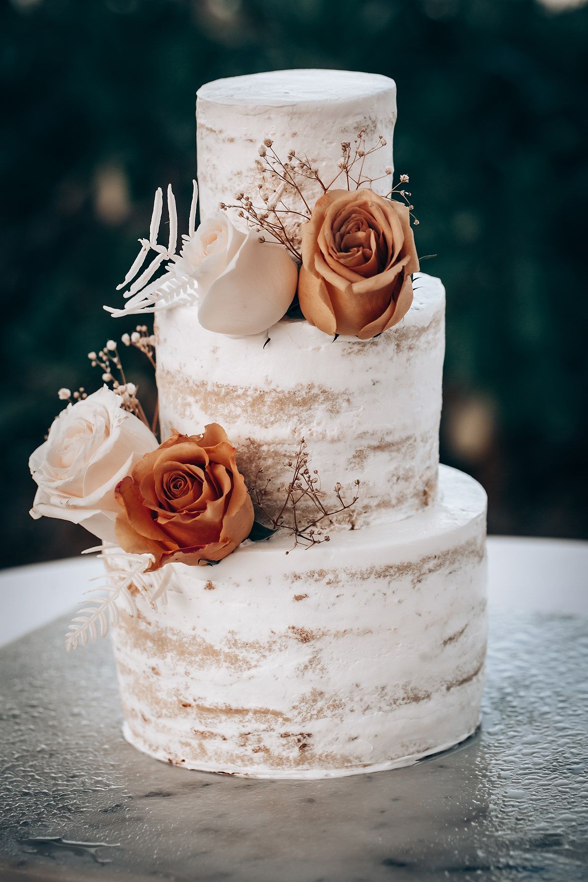 Semi-naked white wedding cake decorated with peach-colored roses and delicate baby's breath on a textured surface.