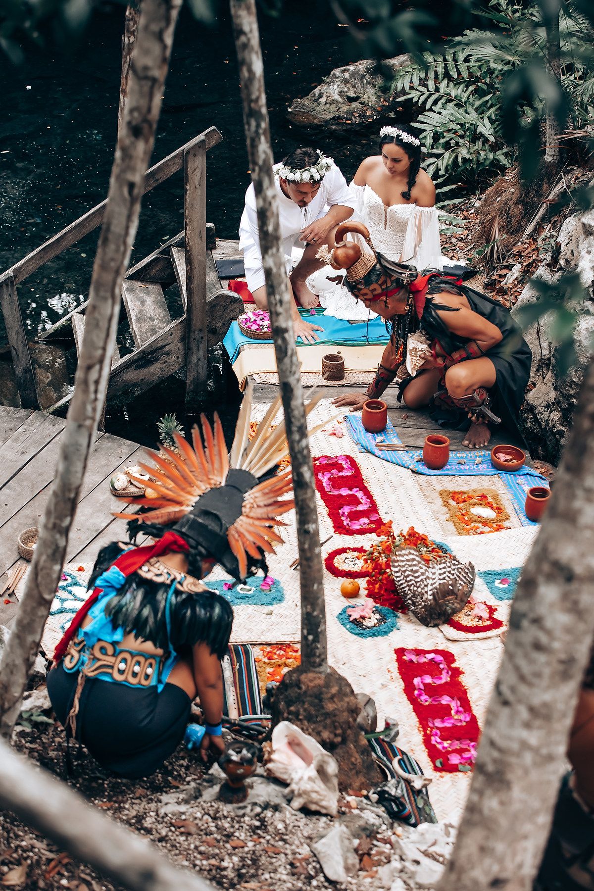A group enjoying a picnic spread with snacks and refreshments laid out on a red and white patterned blanket in a wooded area.