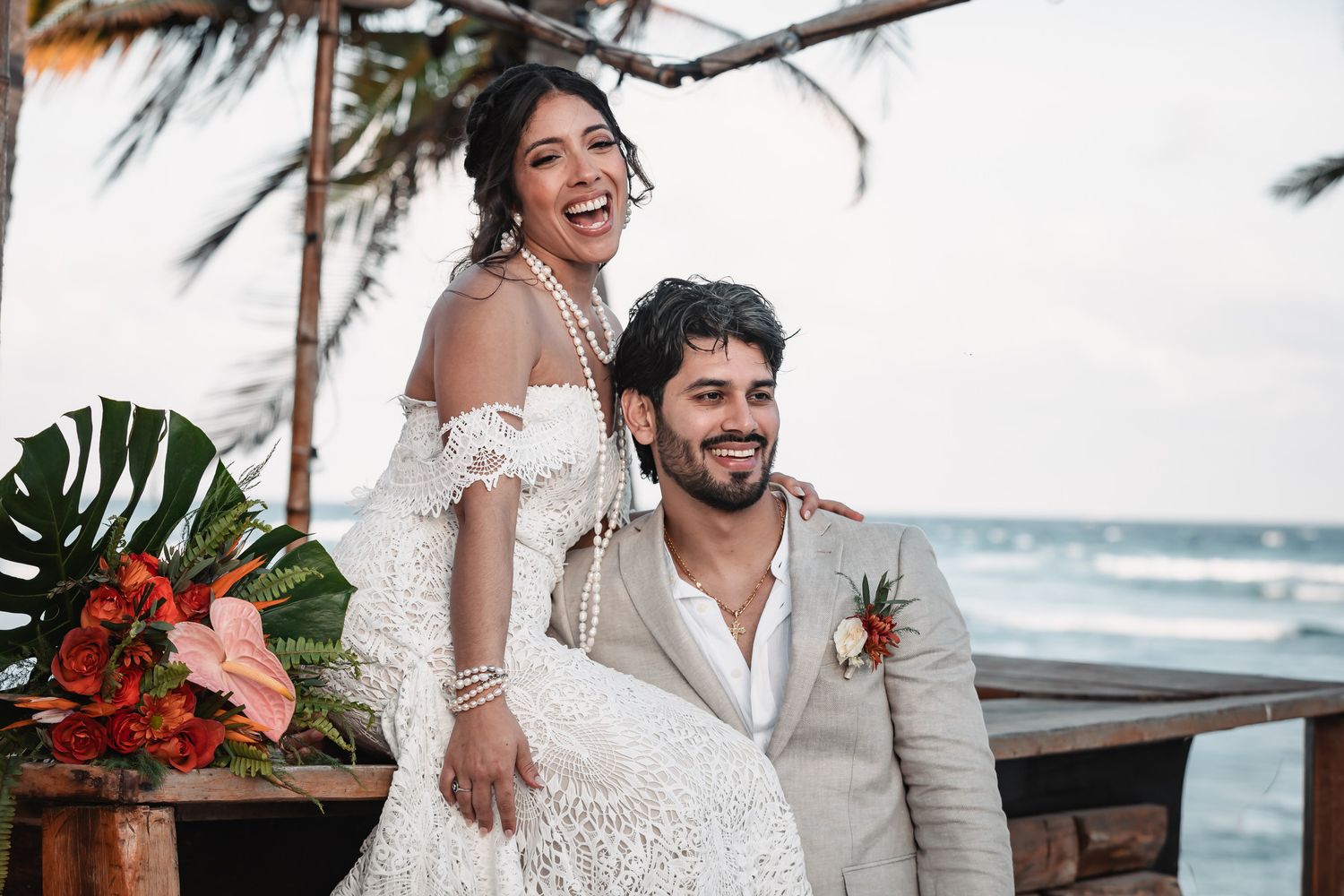 Joyful bride in lace off-shoulder gown and groom in beige suit pose together at oceanside tropical wedding venue.
