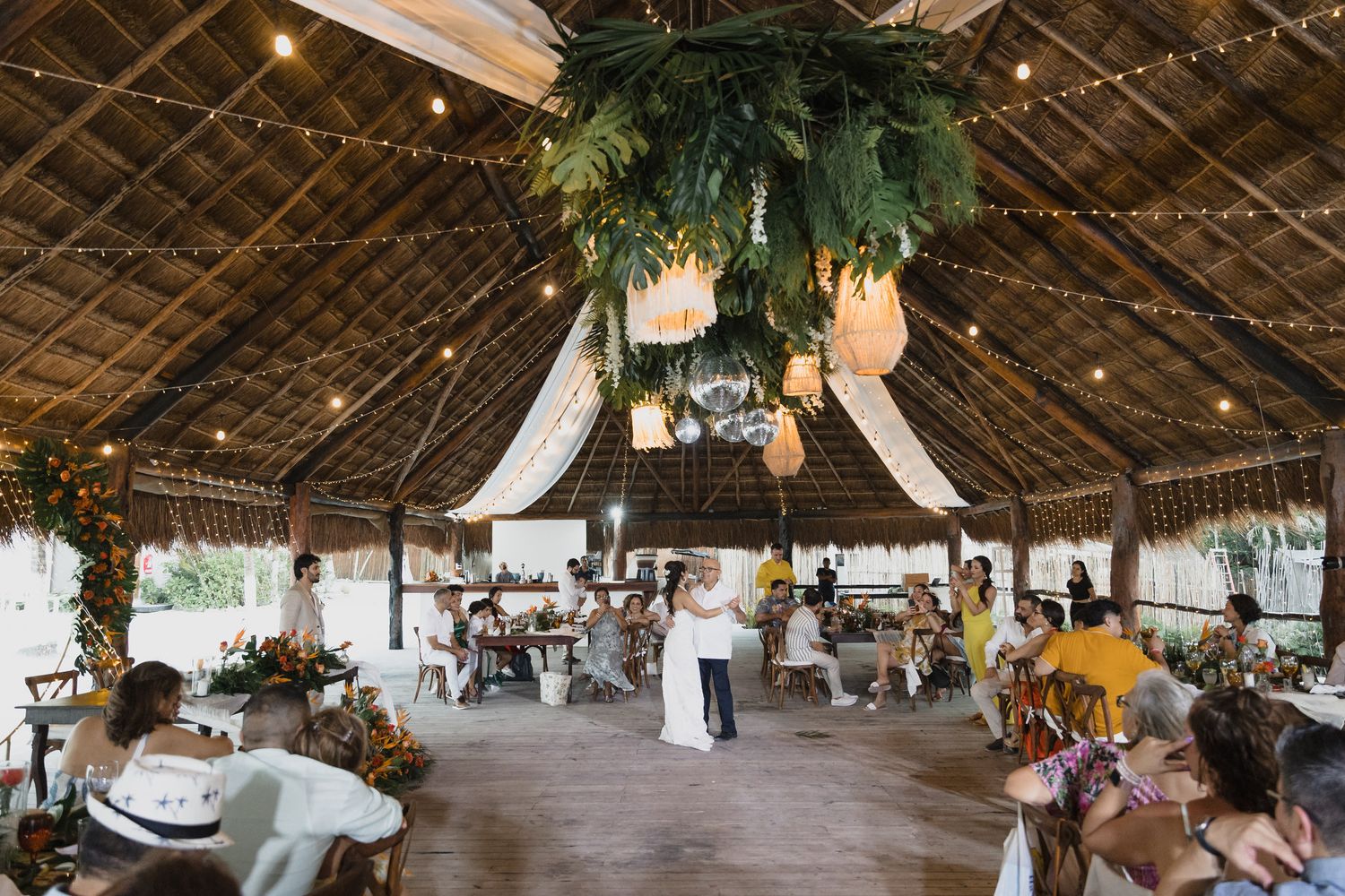 Wedding reception first dance under decorated thatched roof with guests seated around.