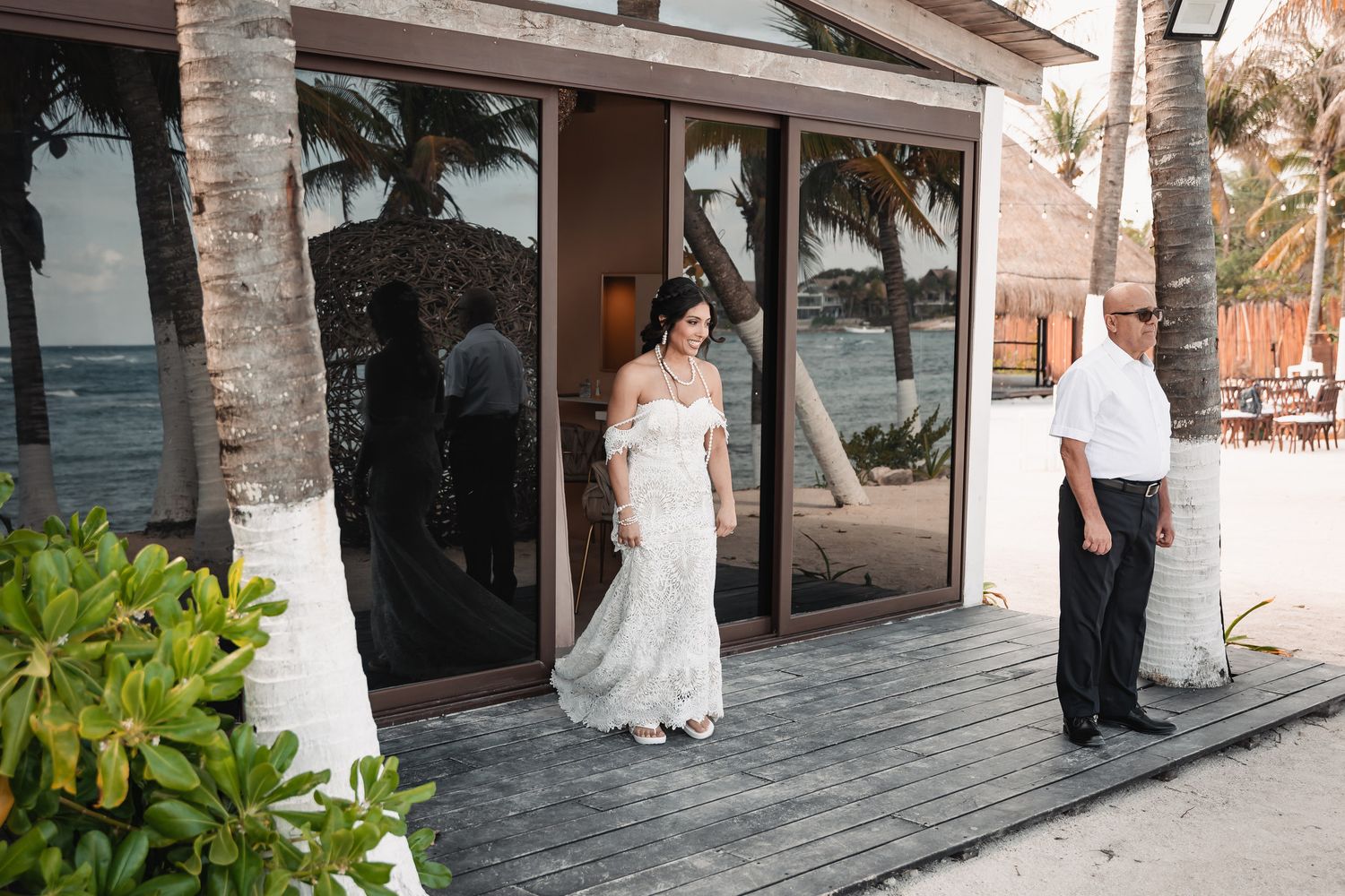 Bride in lace mermaid gown exits beachfront venue doors with escort on tropical wedding day.