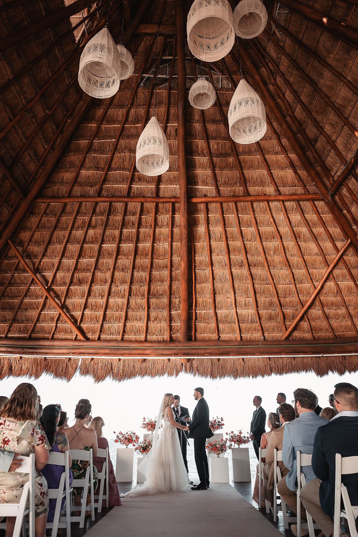 Wedding ceremony setup under a thatched palapa roof with white chairs and floral decorations.
