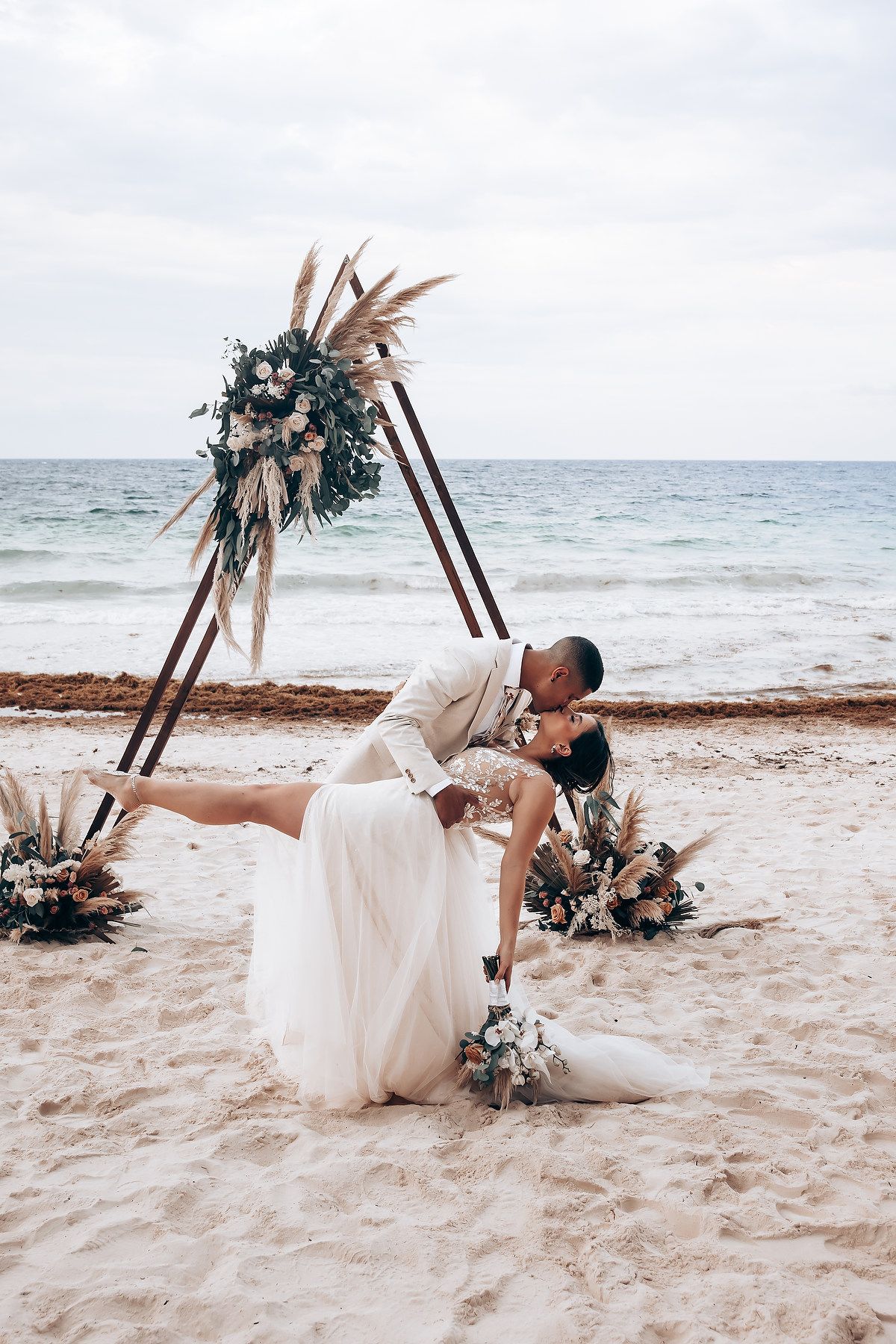 A romantic dip kiss on the beach under a triangular arch decorated with dried flowers.