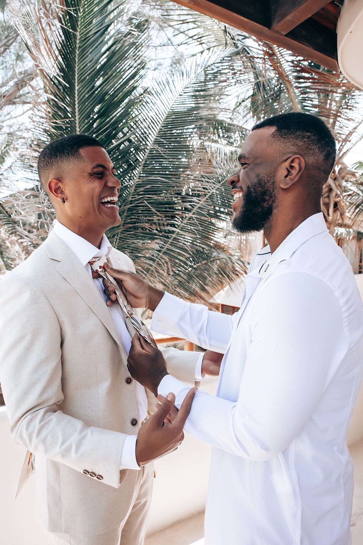 Two men in white and beige formal attire adjust a bowtie against a tropical palm background.