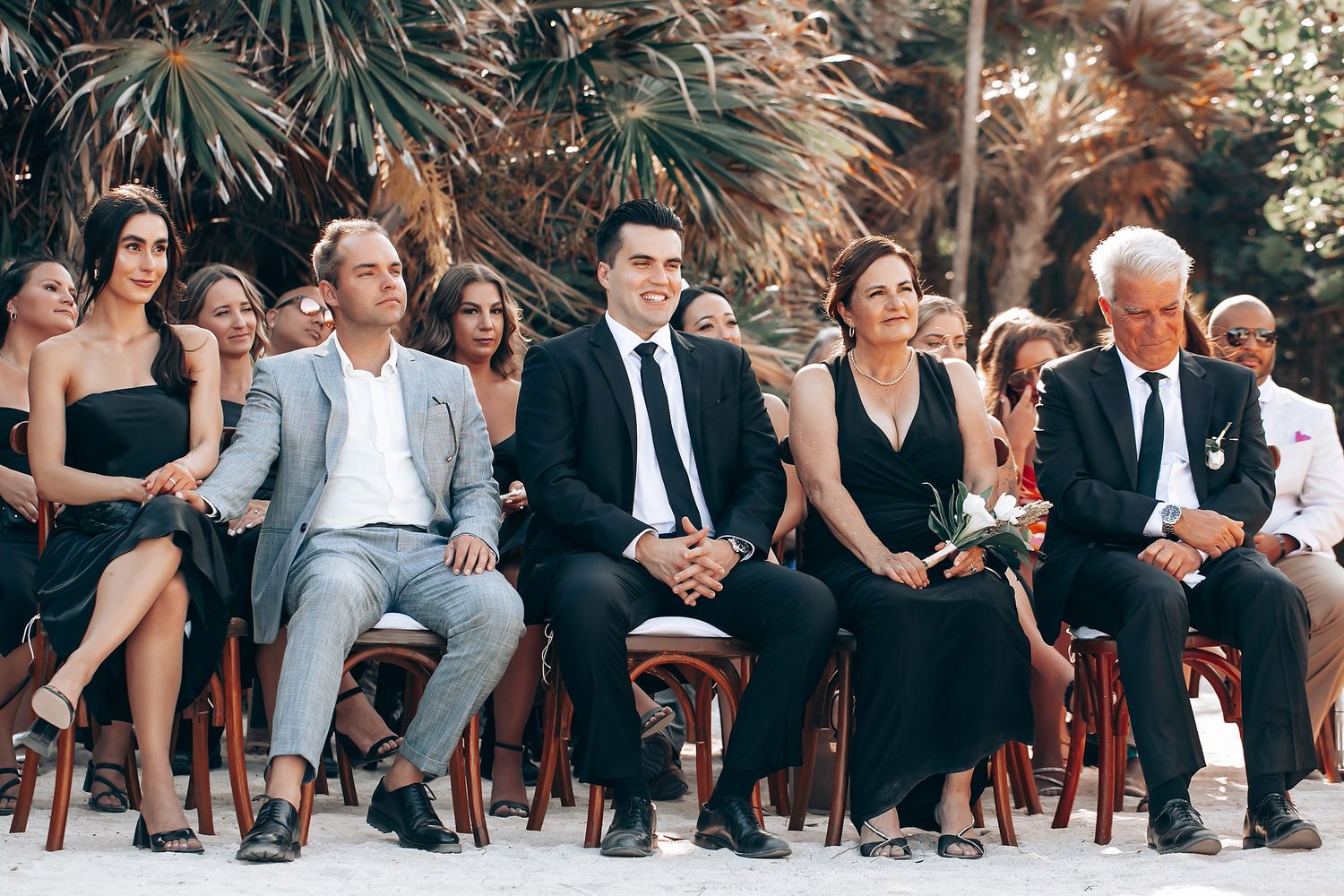 Wedding guests seated in wooden chairs at outdoor tropical ceremony venue during sunset.