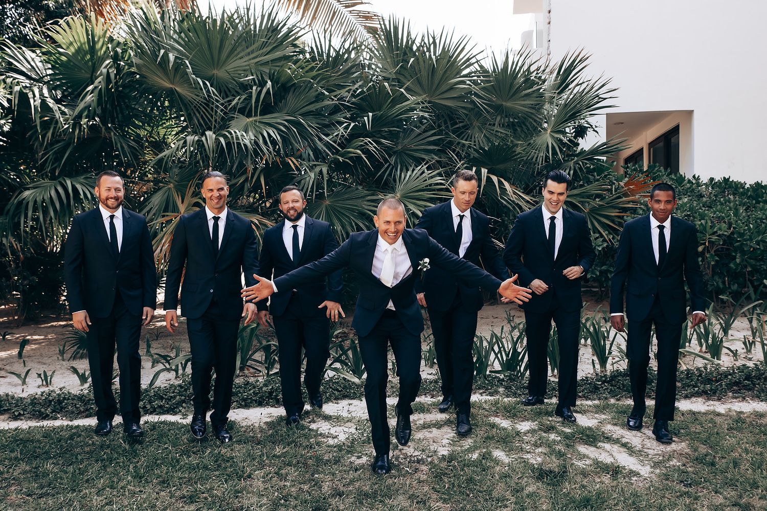 Wedding party in formal attire posing outdoors with palm trees in background.
