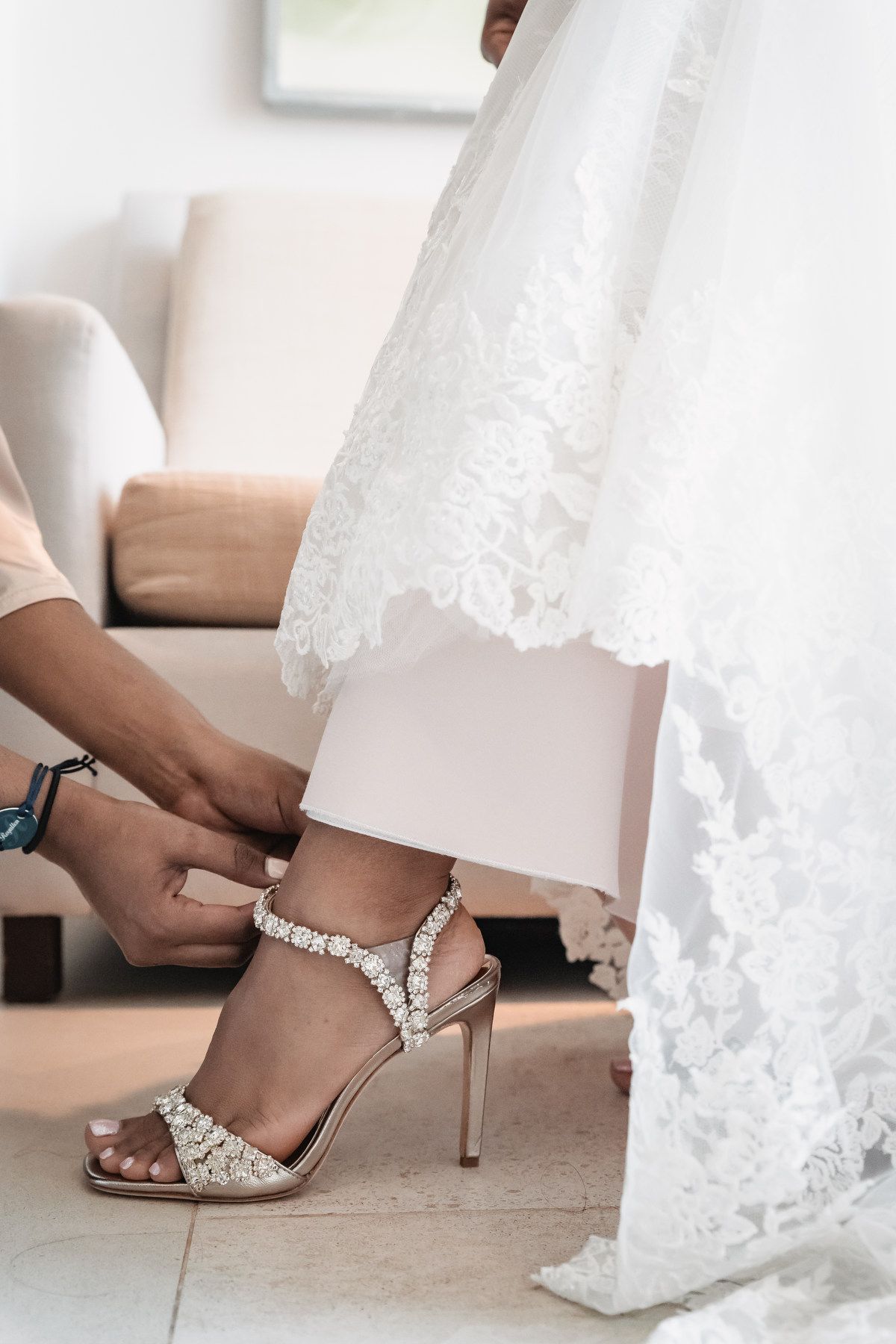 Helper fastening a crystal-embellished strappy heel on a bride in a lace wedding gown.