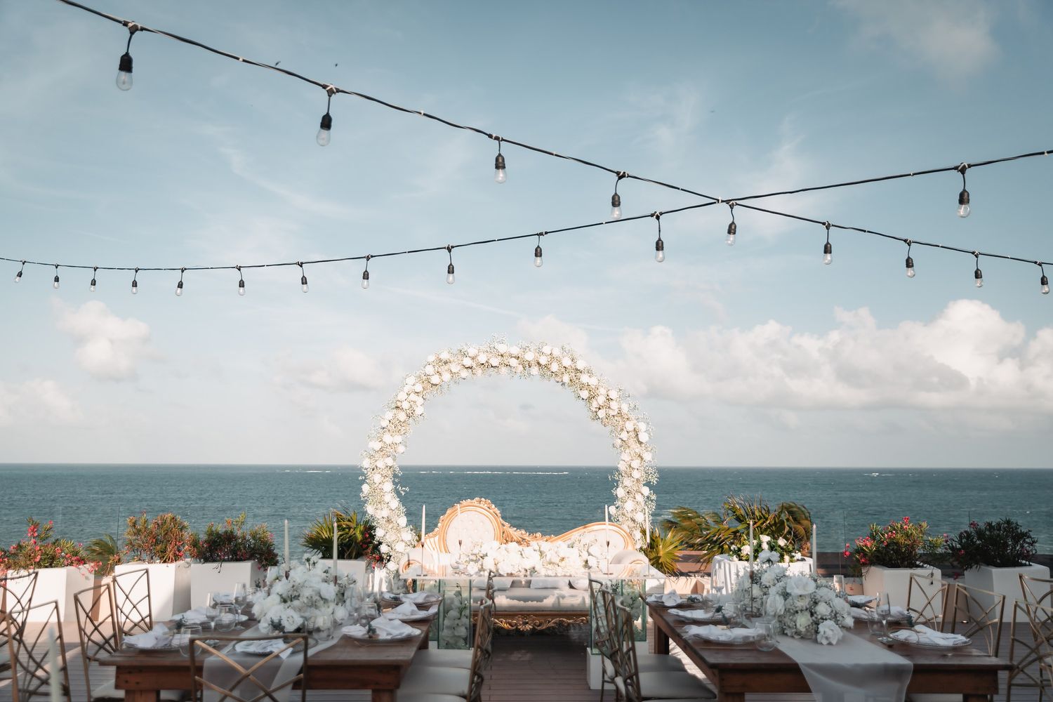 Outdoor beachfront wedding reception with a floral arch and string lights over the ocean.