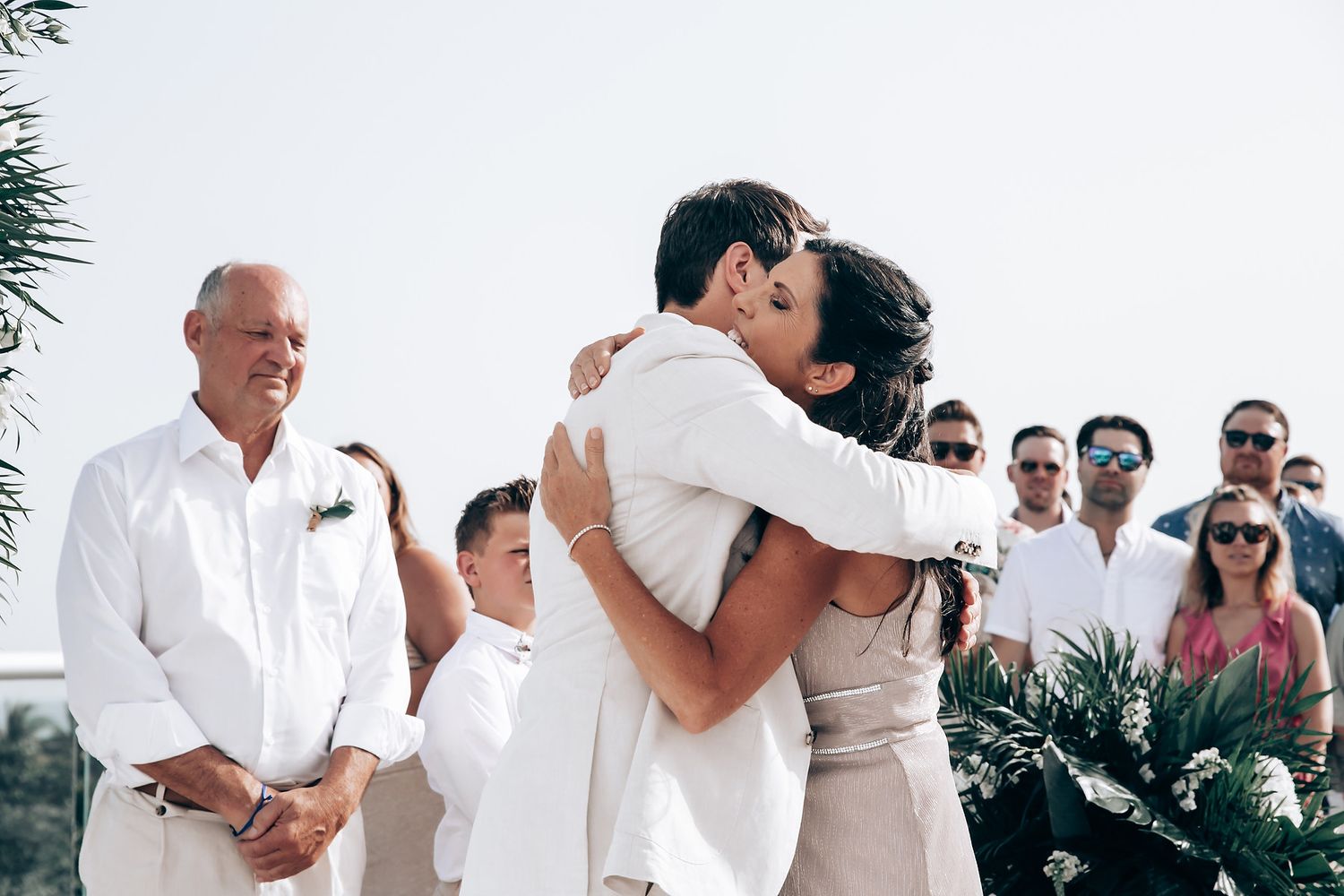 Intimate outdoor wedding ceremony with guests watching couple embrace under tropical foliage.