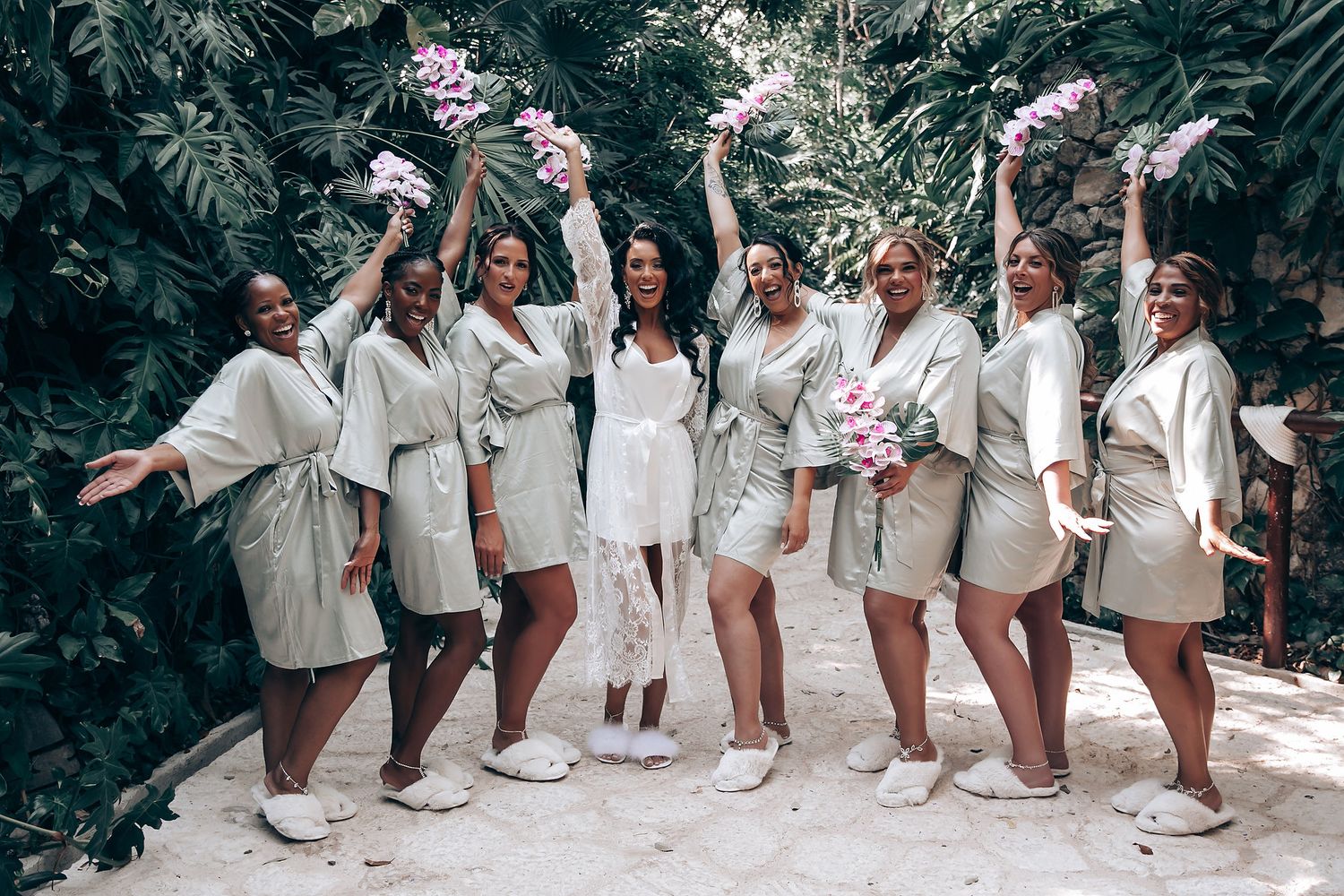 Wedding party members in white attire pose together in a tropical garden setting.