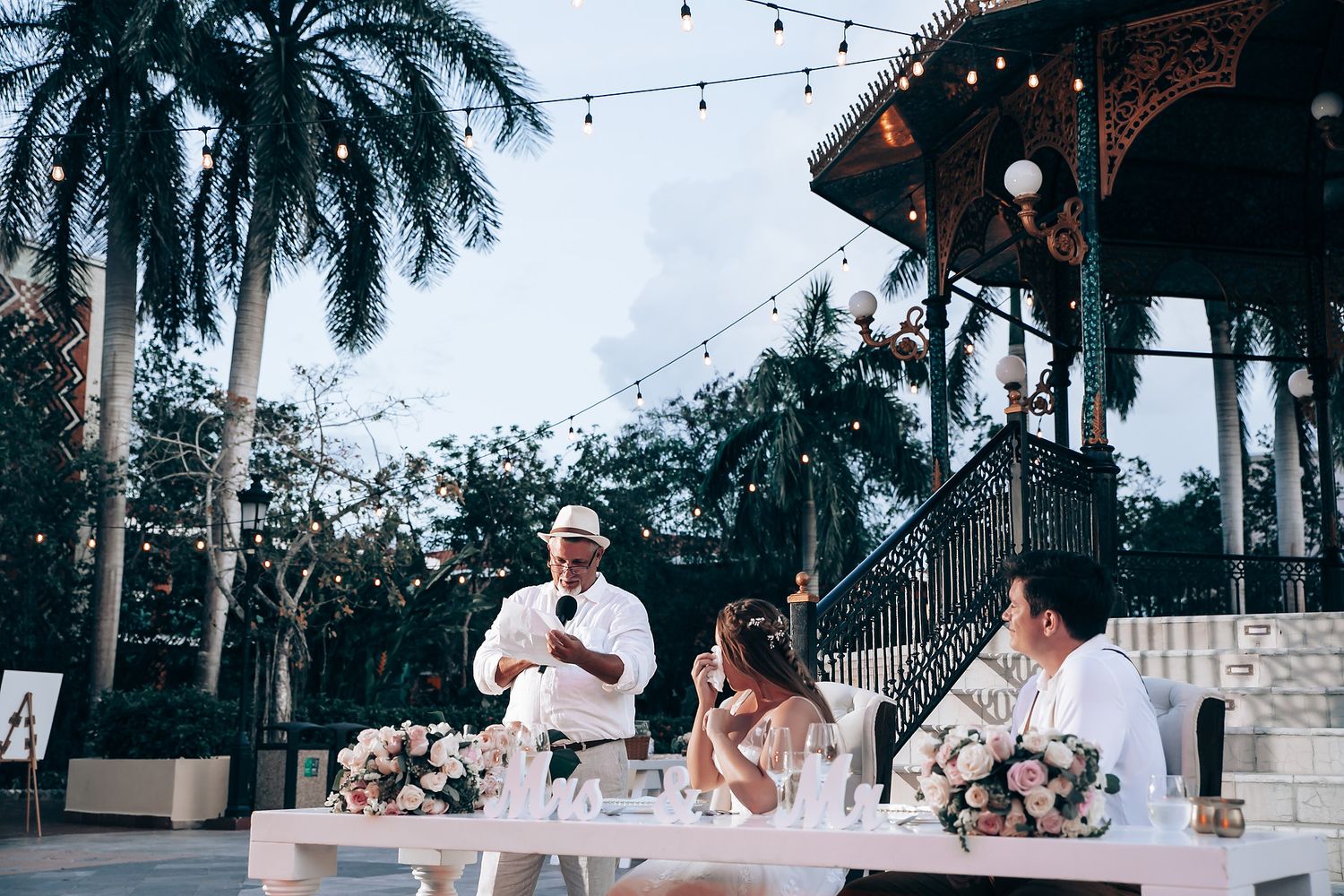Evening wedding reception under string lights with palm trees in background.