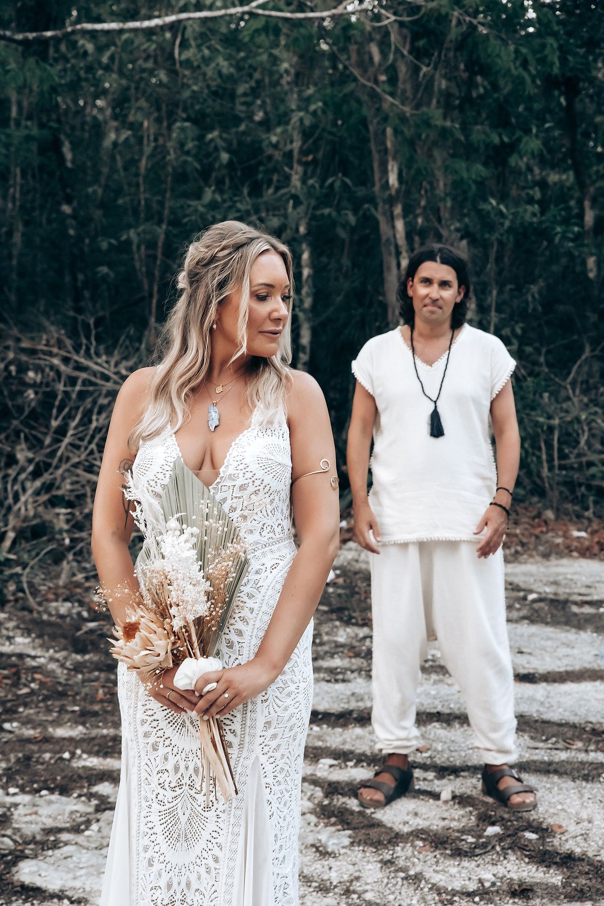 Two people in white dresses stand in a rustic outdoor setting holding dried floral arrangements.