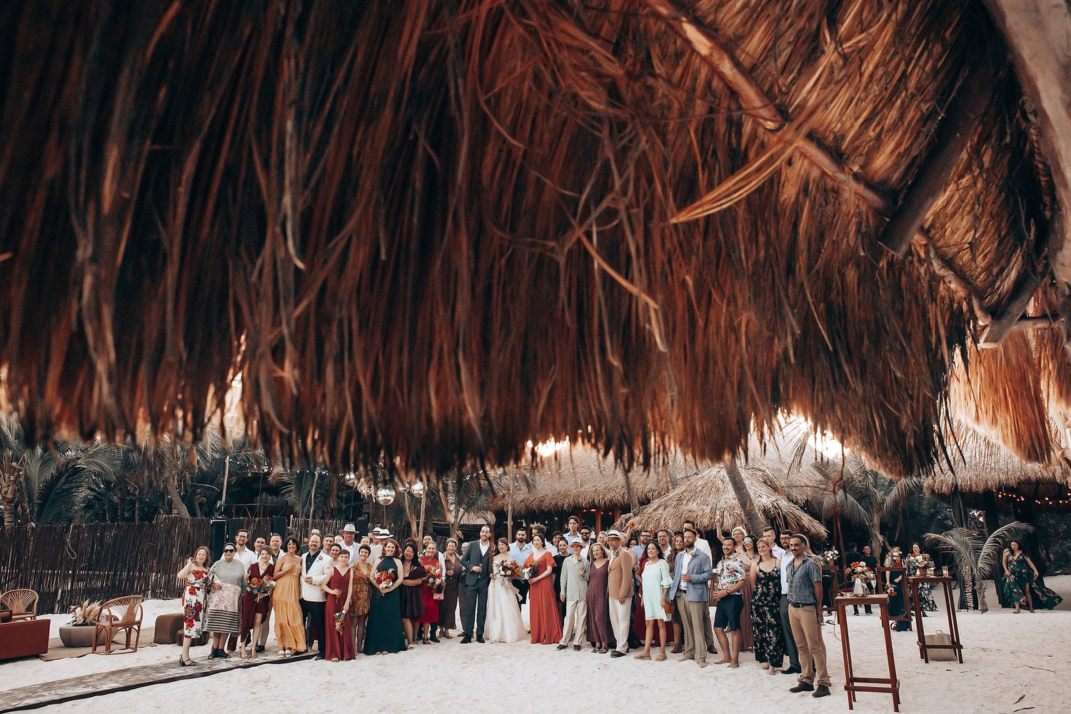 Wedding ceremony setup under a thatched palm roof with rows of guests seated on wooden chairs.