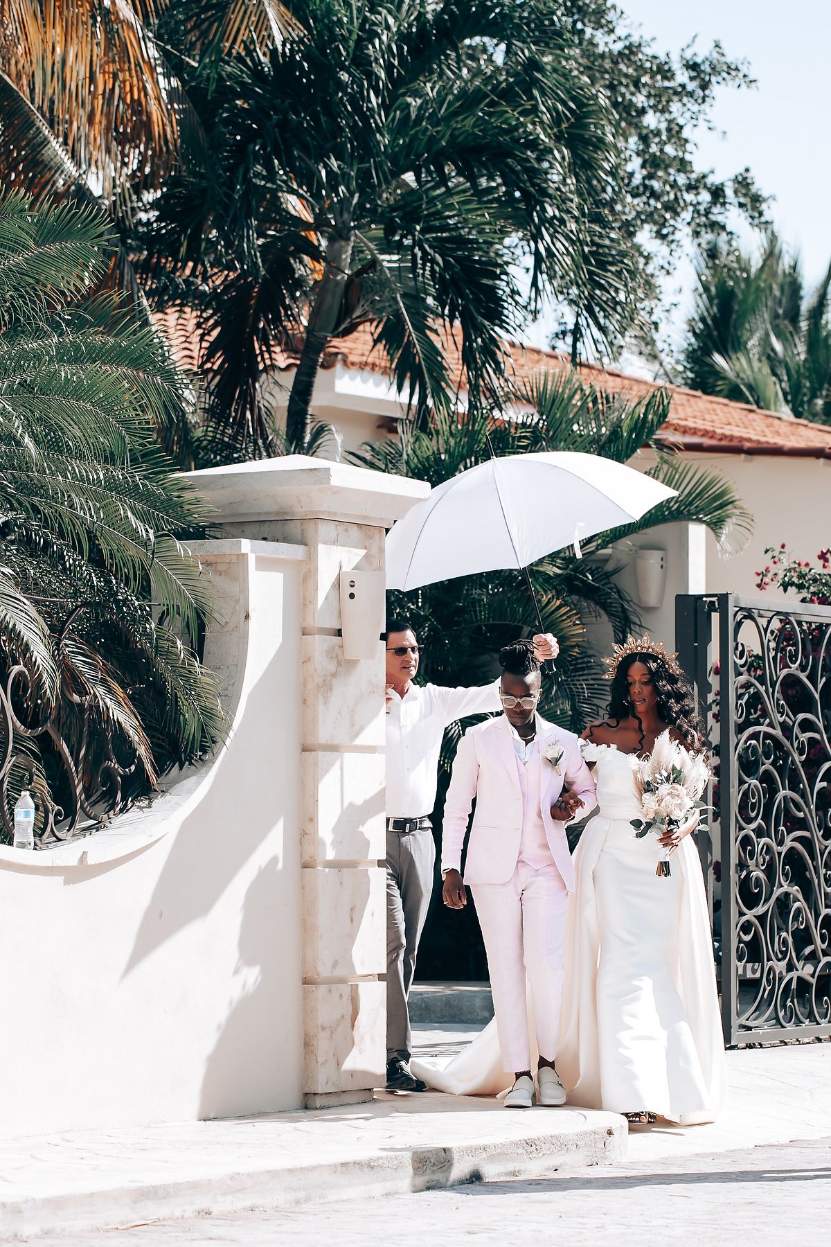Wedding party in white attire poses elegantly against a Mediterranean-style white wall with palm trees.
