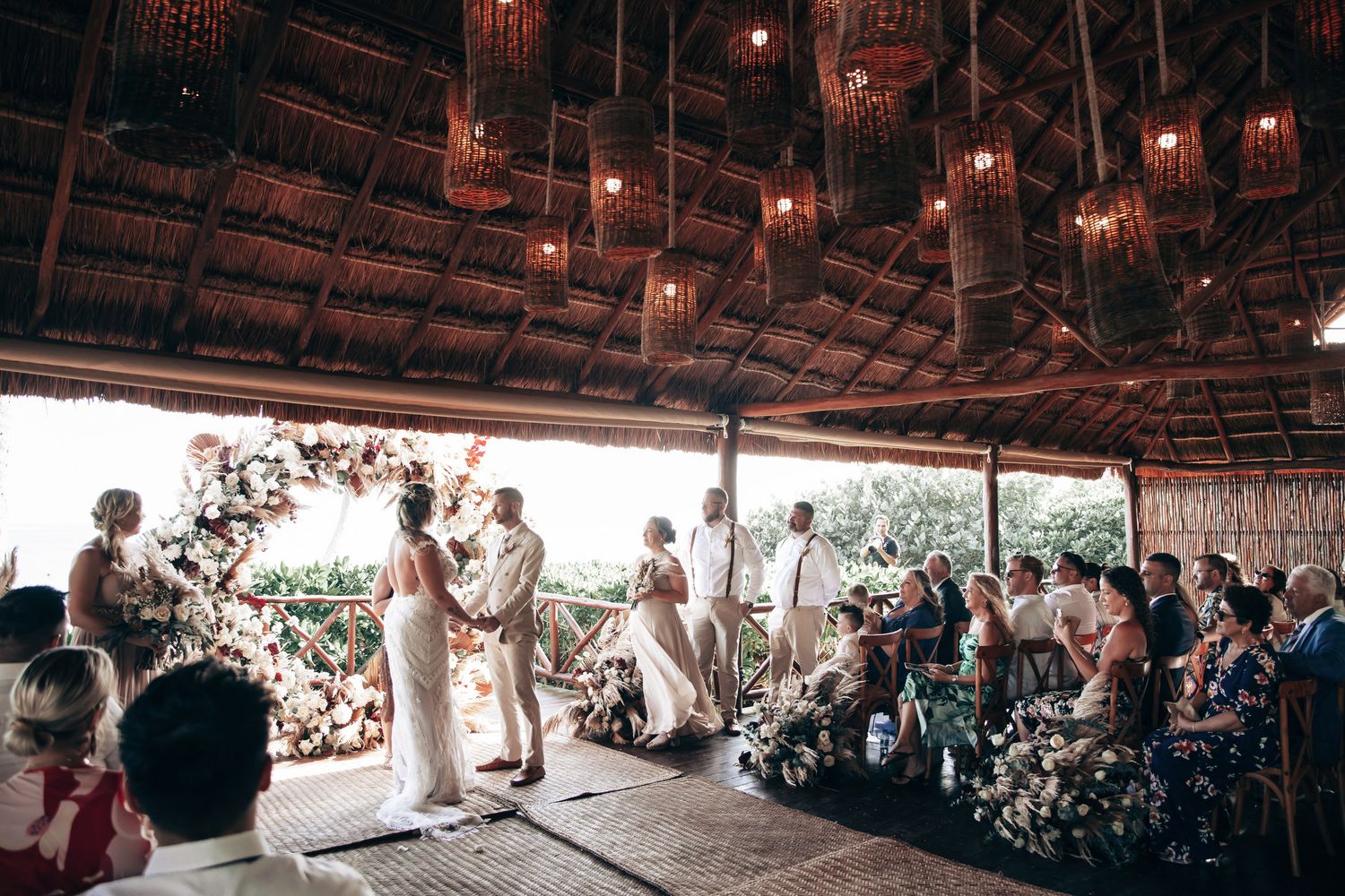 Outdoor wedding ceremony under thatched roof with hanging lanterns, floral arch, and seated guests in tropical venue at Dreams Tulum