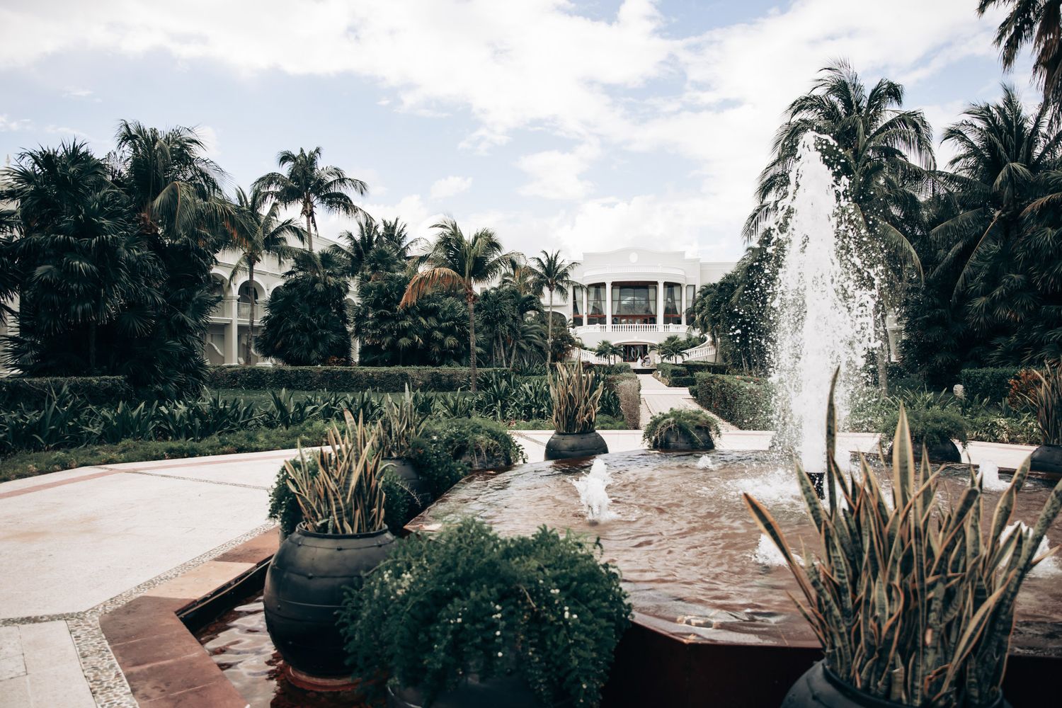 Tropical resort garden with water fountains, lush palm trees, and manicured plants under a cloudy sky.