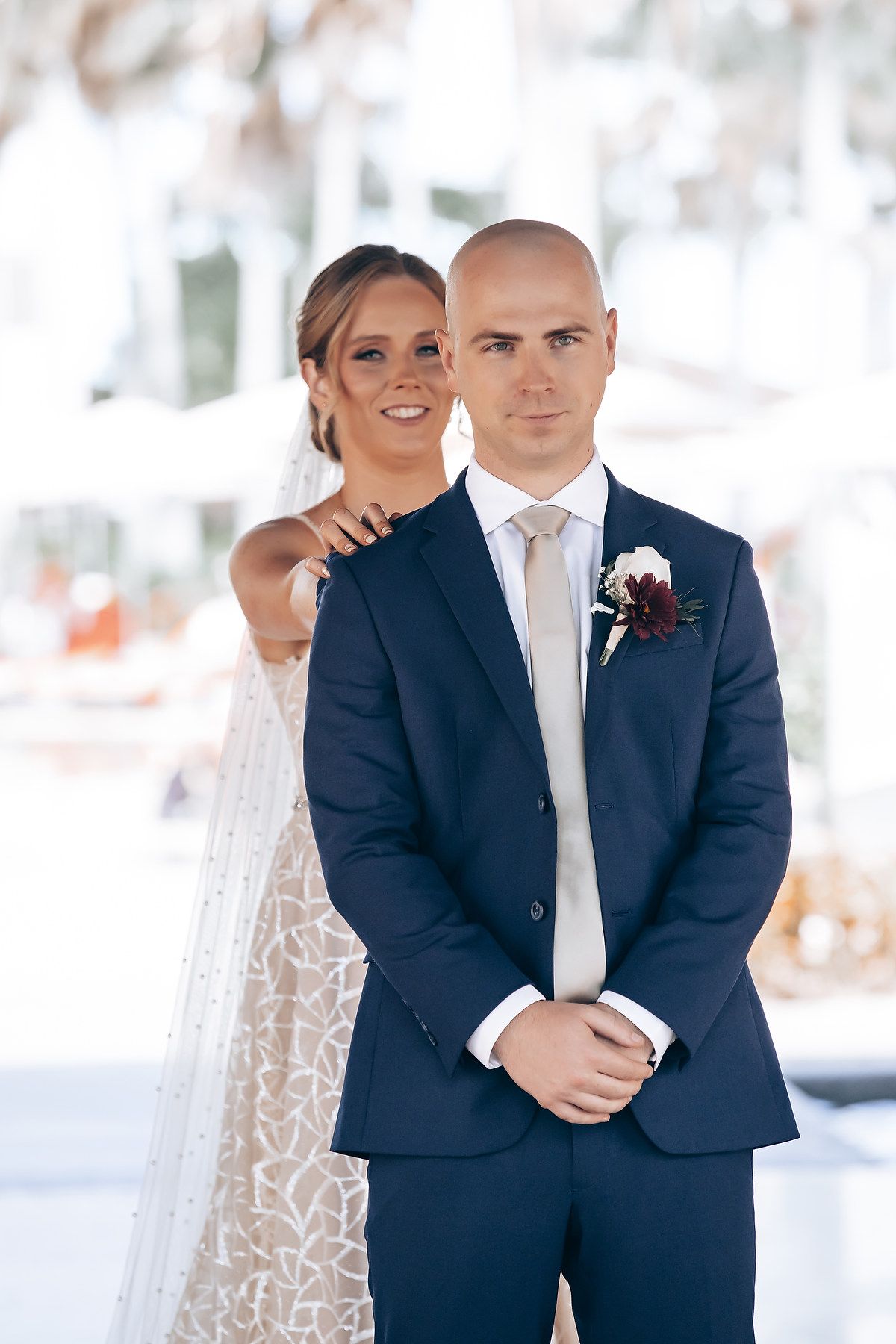 A bride and groom pose for a wedding photo, with the bride wearing a lace dress and the groom in a dark suit.