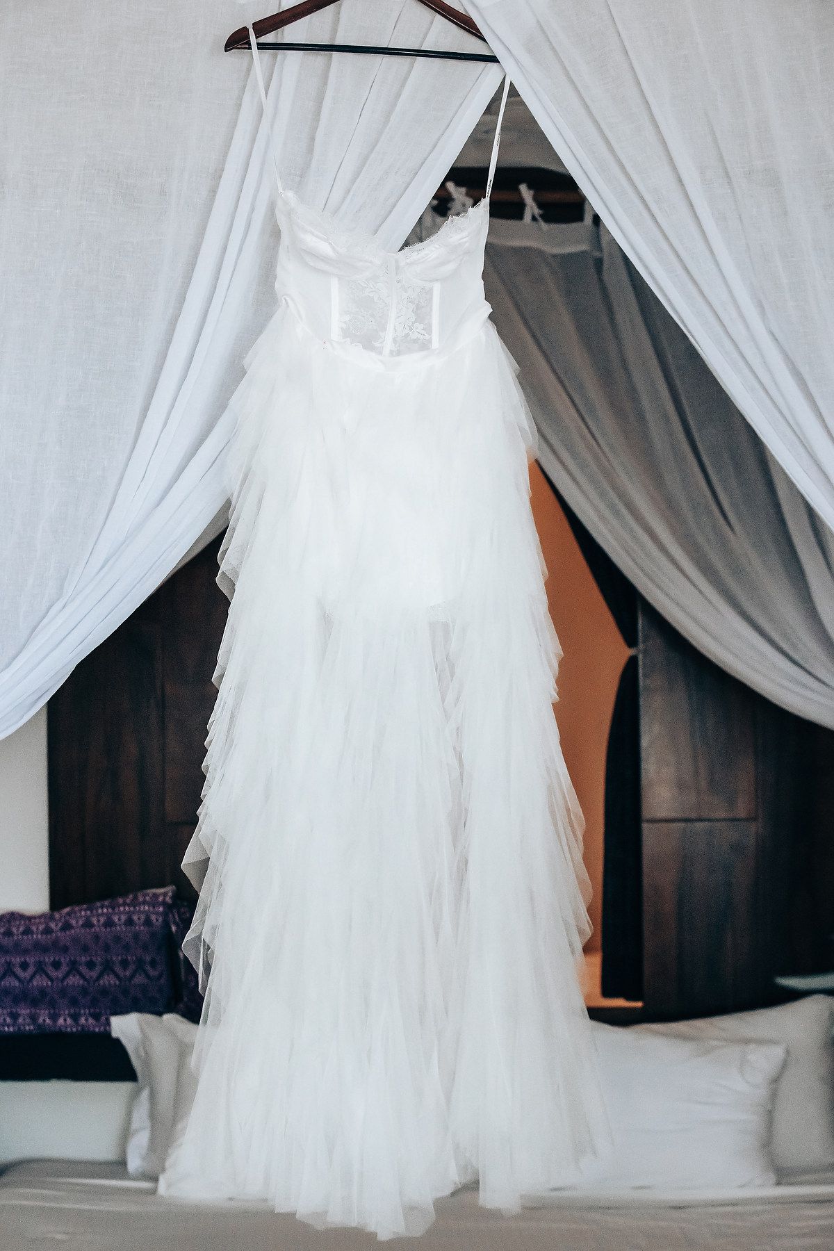 A white wedding dress hangs in a room, with sheer curtains and a wooden hanger.