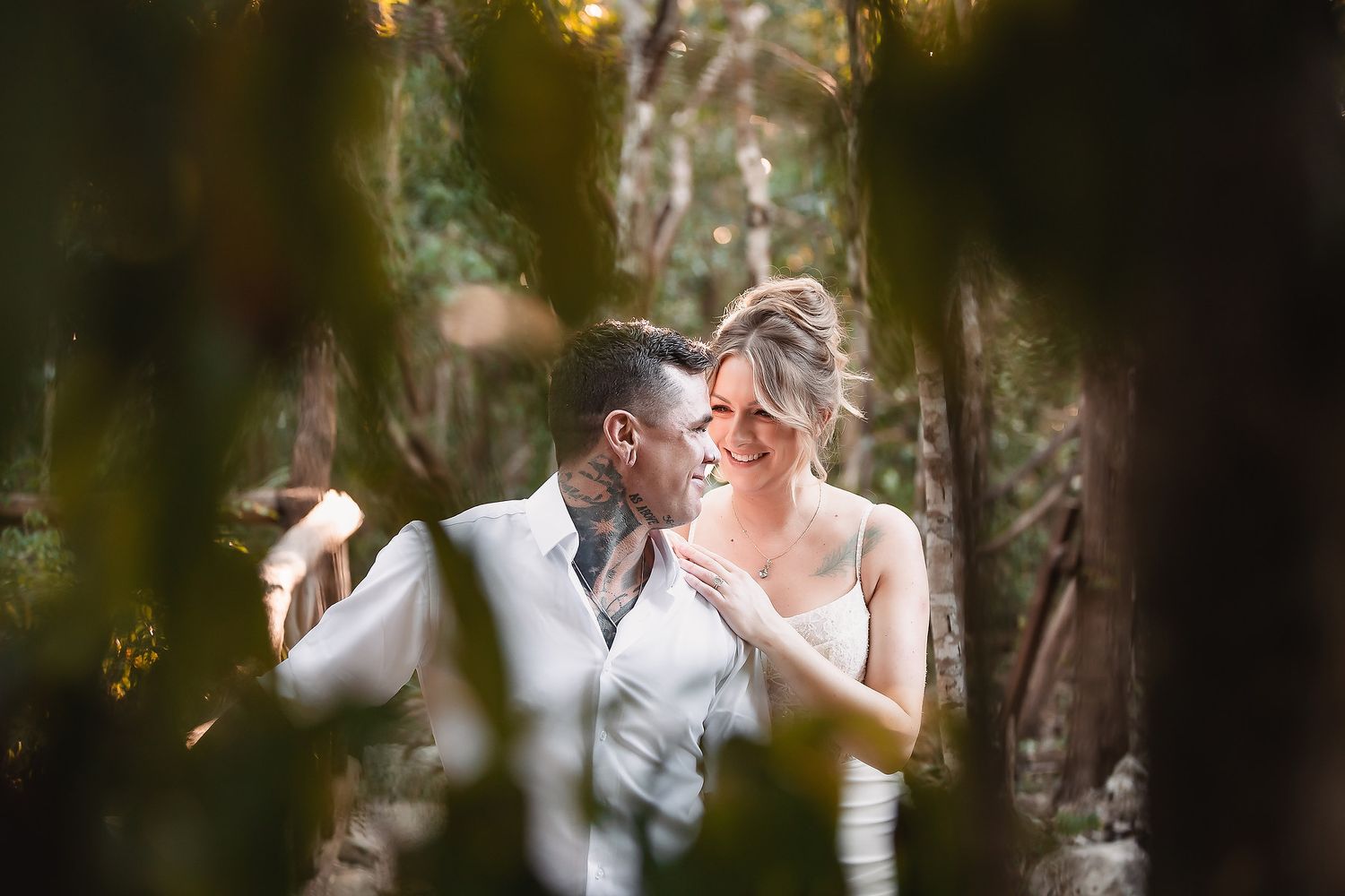 A couple shares a romantic moment surrounded by lush greenery, the bride in white and groom in a suit with tattoos.