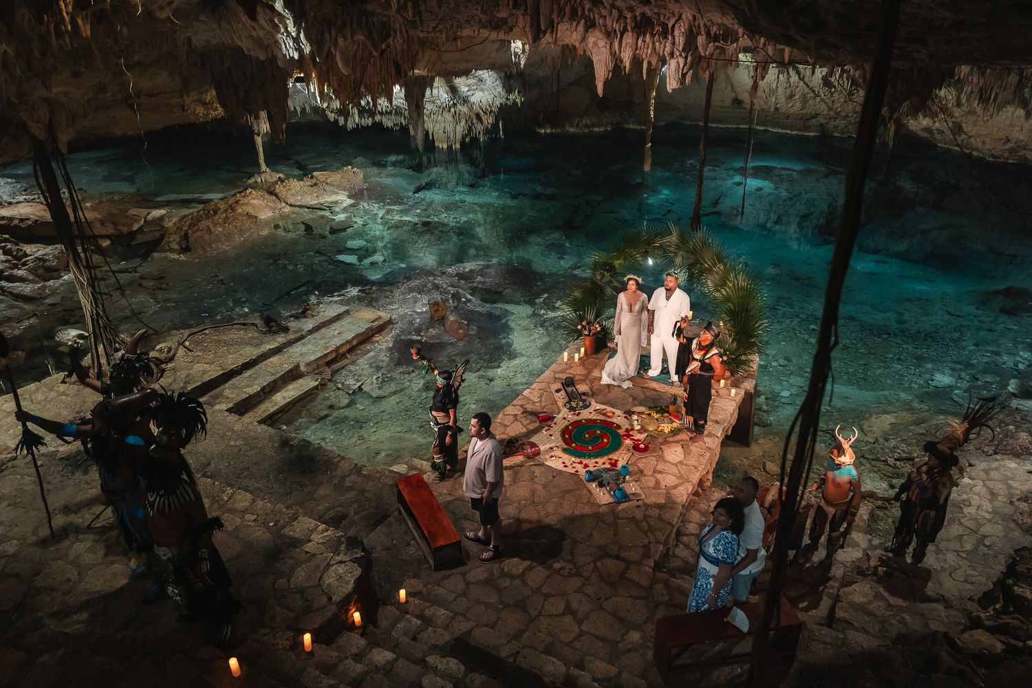 Aerial view of a mystical cenote wedding ceremony with candles, lush greenery, and turquoise water in a cave.