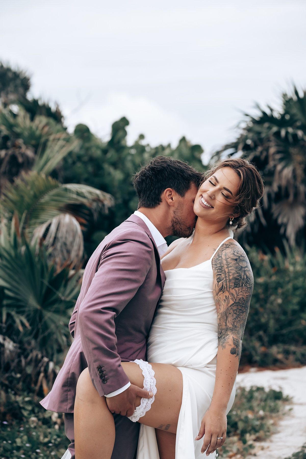 Romantic beach wedding photoshoot with couple in white dress and pink suit among palm trees.