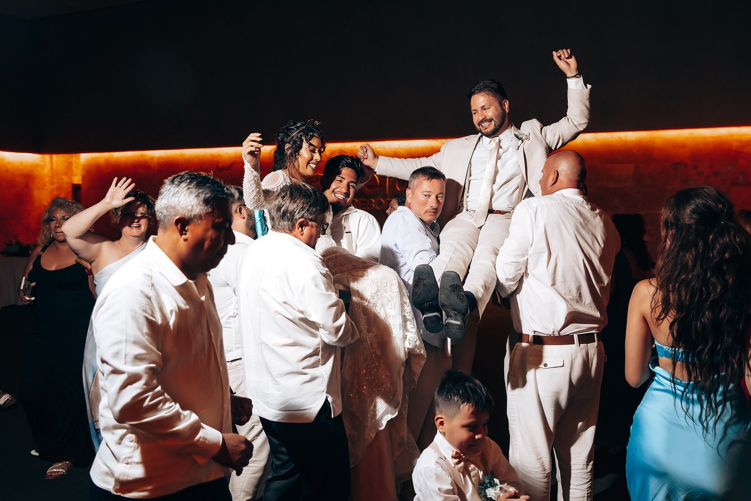 Wedding reception dance party with guests in white attire under warm lighting.
