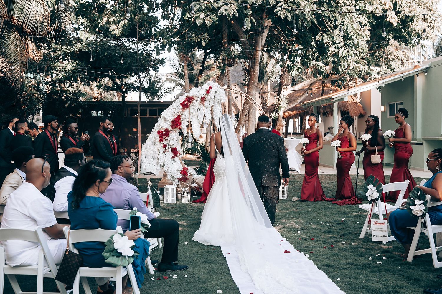 Wedding guests gather outdoors under trees for a celebration with white chairs and decorative elements.