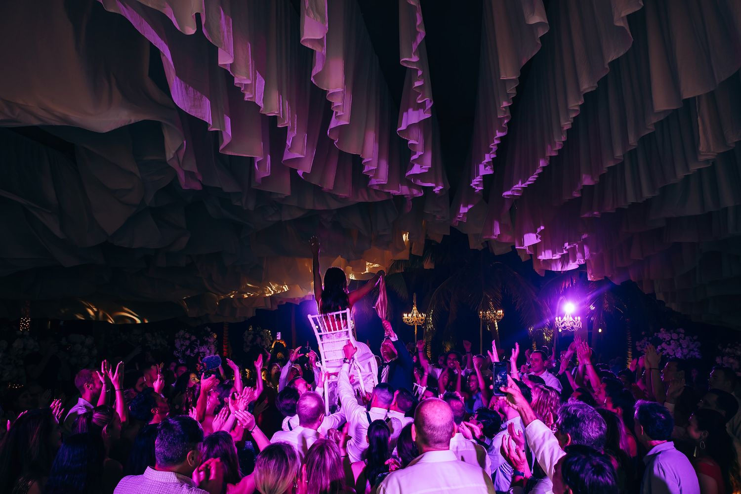 Wedding reception dance floor illuminated with purple lighting and hanging fabric decor.