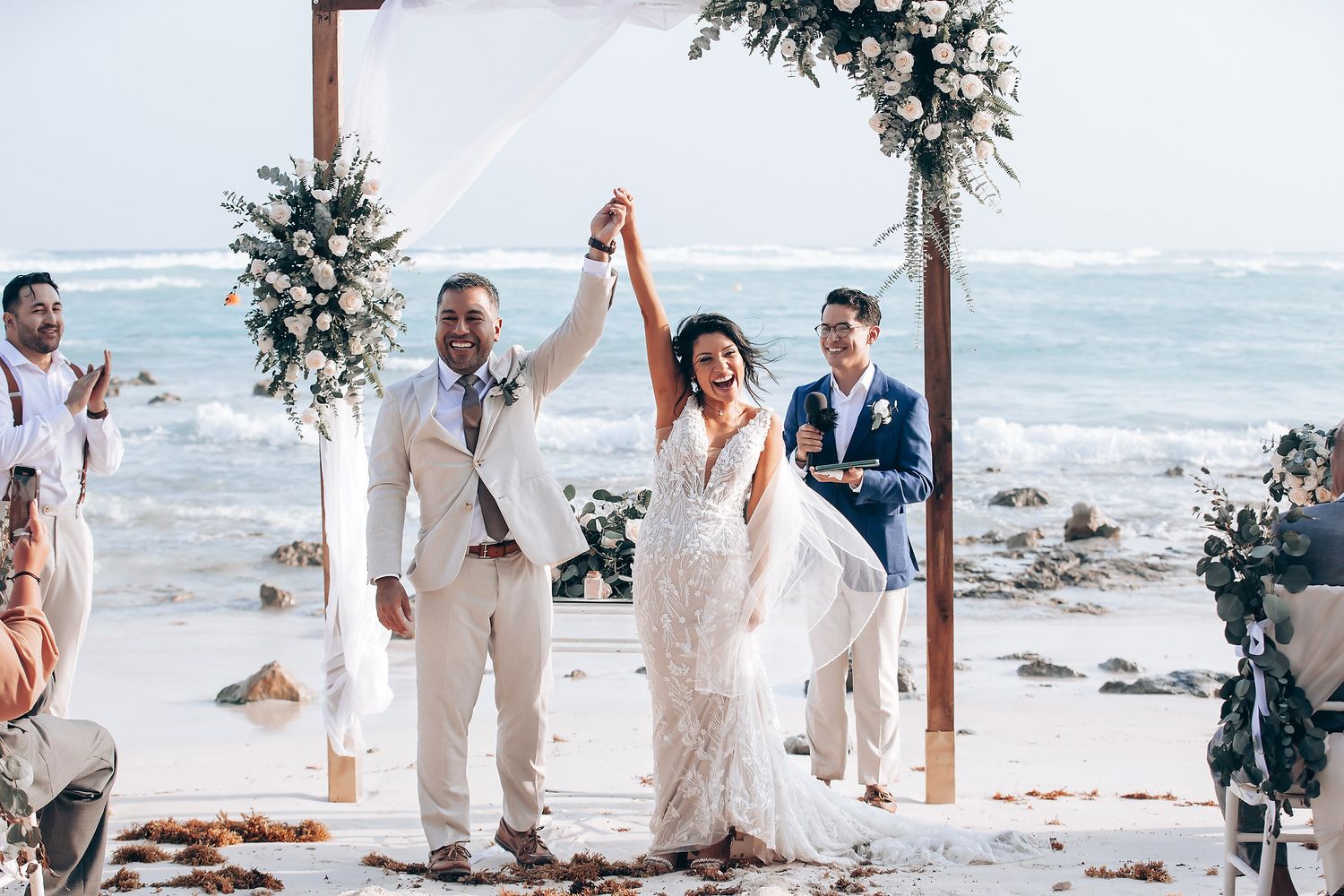 A beachfront wedding ceremony setup with white decor and palm trees along a tropical shoreline.
