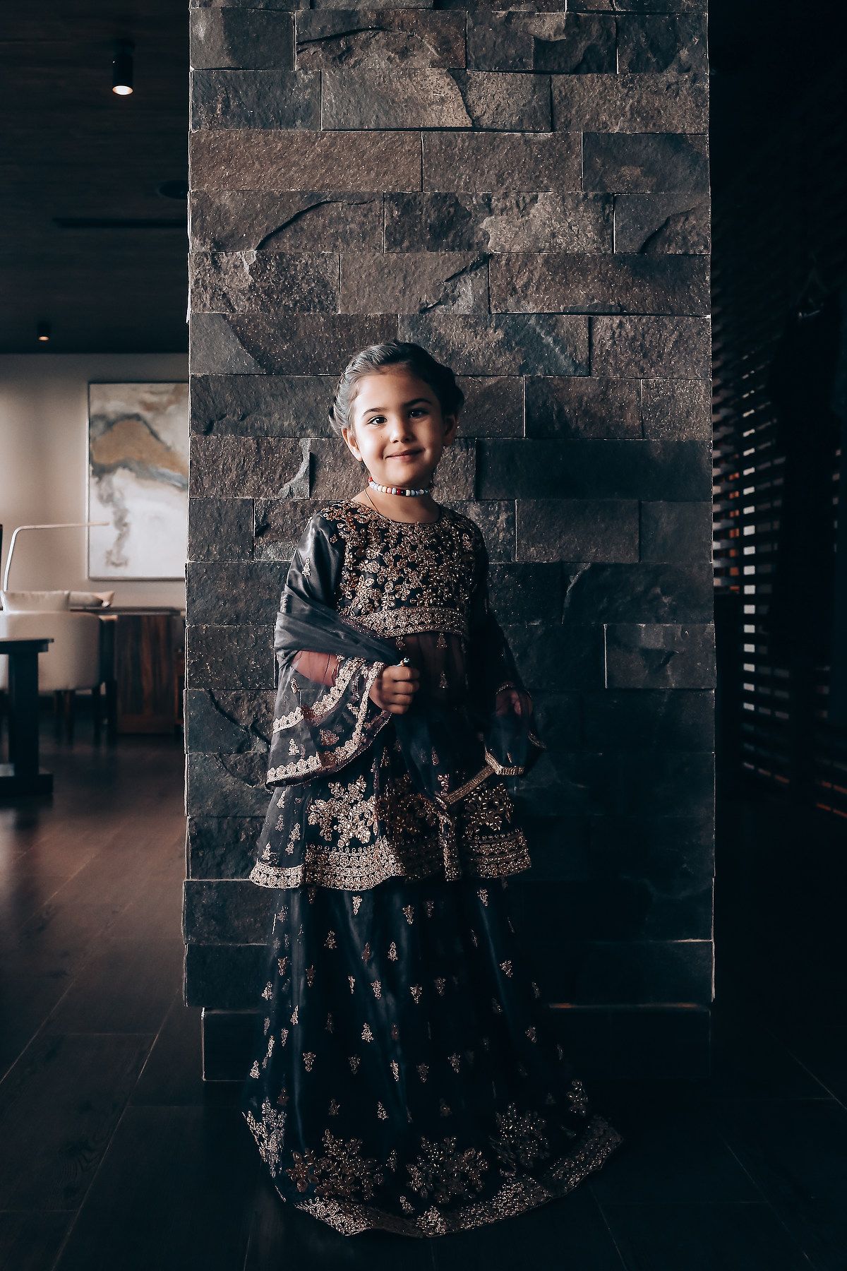 A small child wearing an ornate black and gold traditional Indian lehenga stands against a textured stone wall.