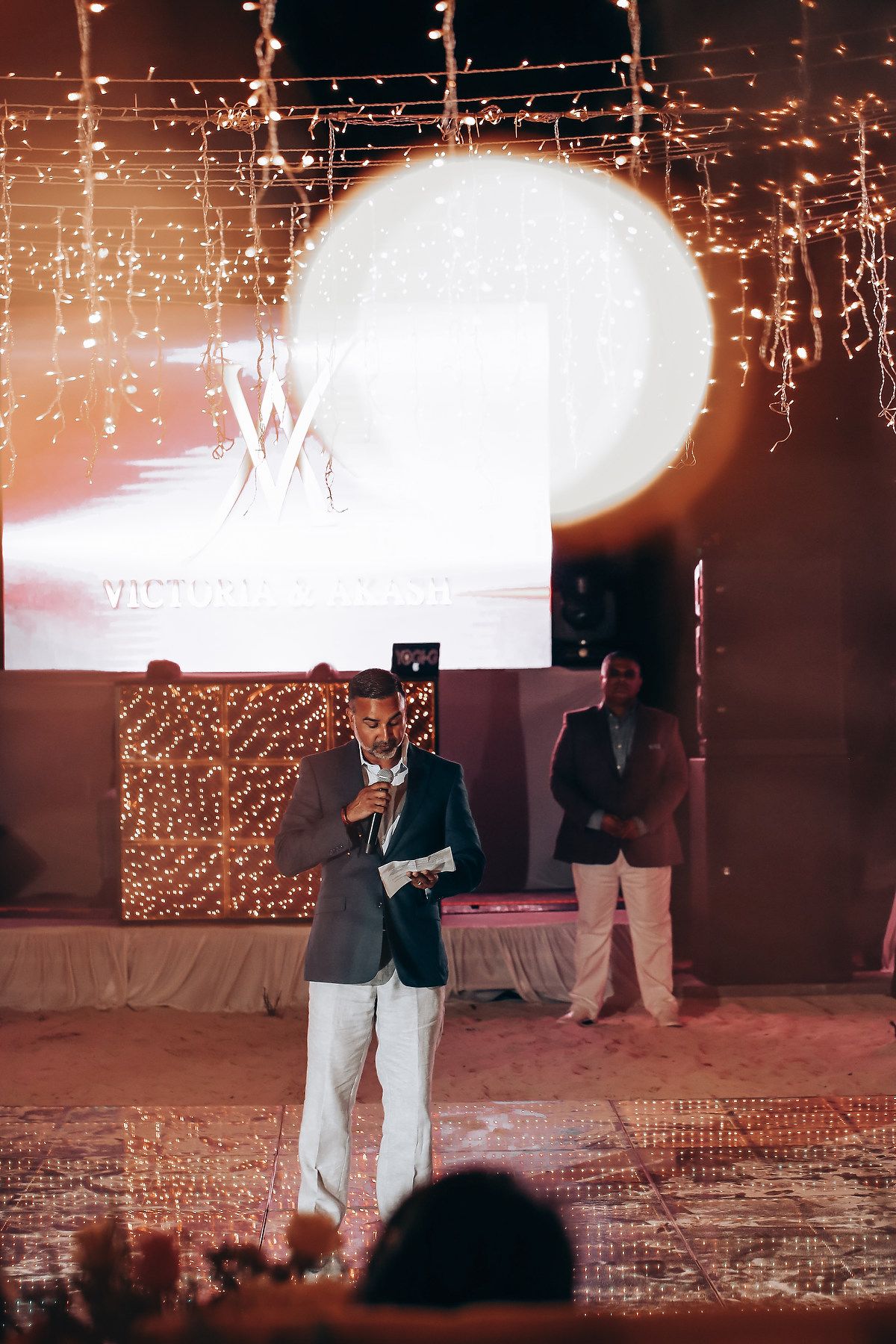A person in a suit stands on stage with dramatic lighting and sparkly effects in the background at an event.