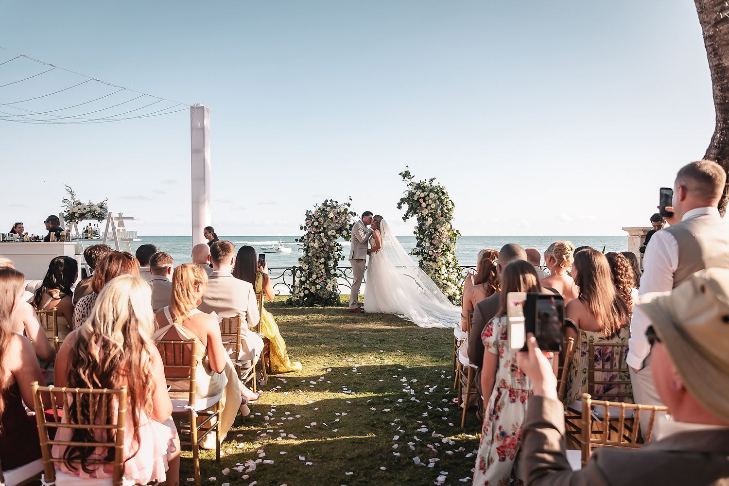 Wedding guests seated on white chairs watch ceremony by the ocean under swaying palm trees.