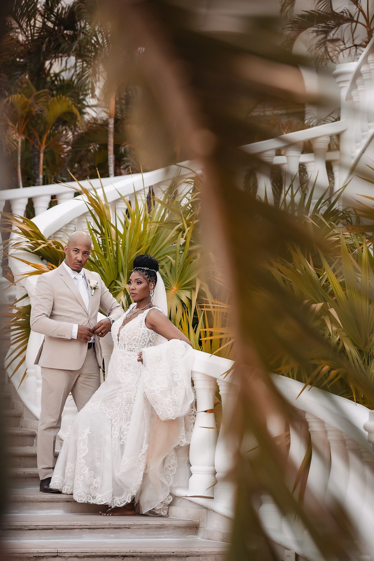 Couple in white attire sitting on grand outdoor staircase with ornate white railings and palm trees.