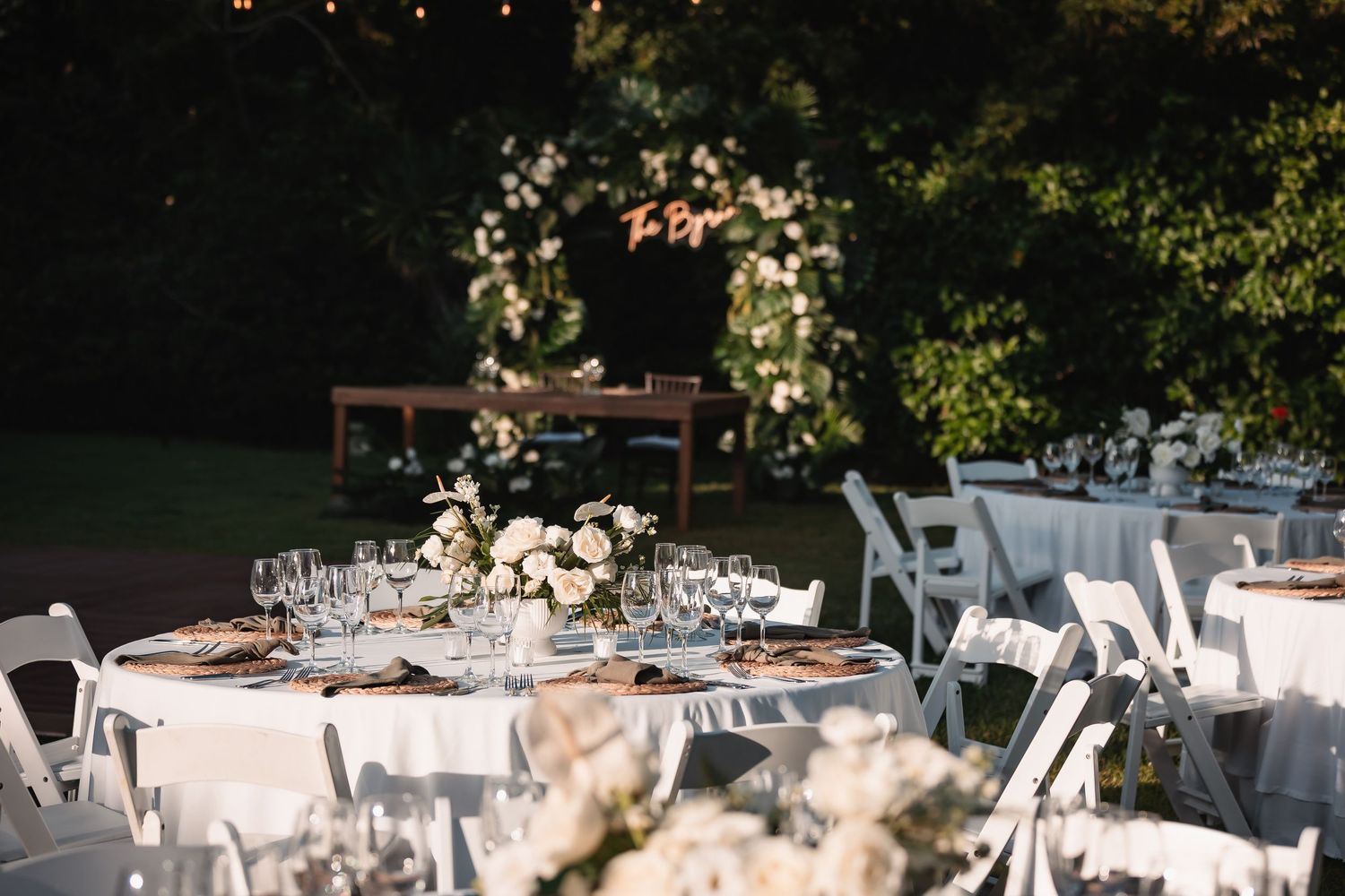 Wedding reception tables decorated with white linens and floral arrangements under palm trees.