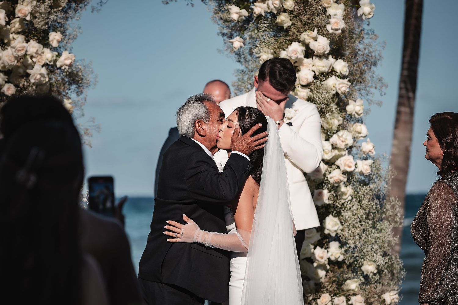 Emotional wedding moment under a floral arch with ocean backdrop during sunset ceremony.