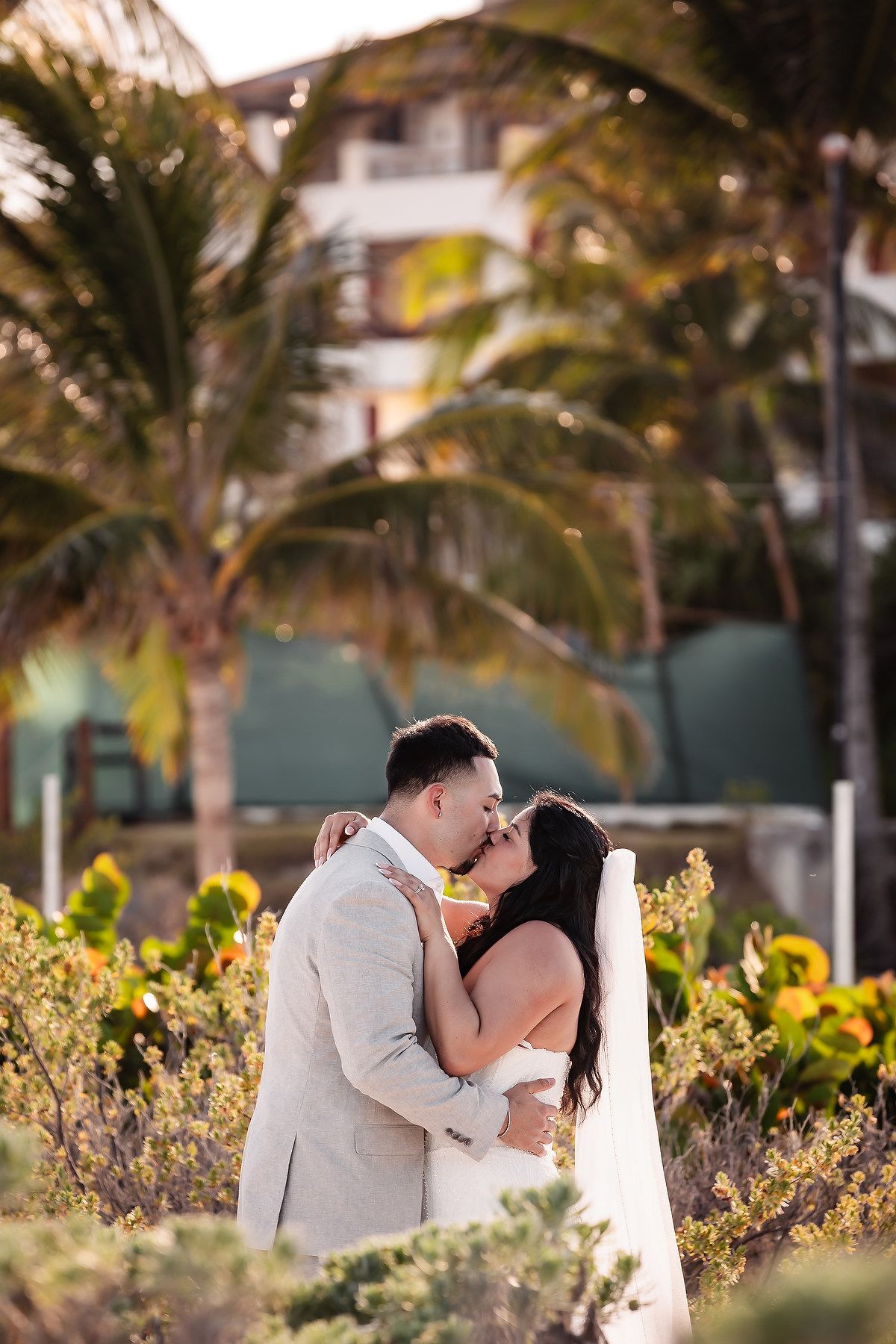 A romantic wedding moment captured among tropical palm trees and yellow flowers at sunset.