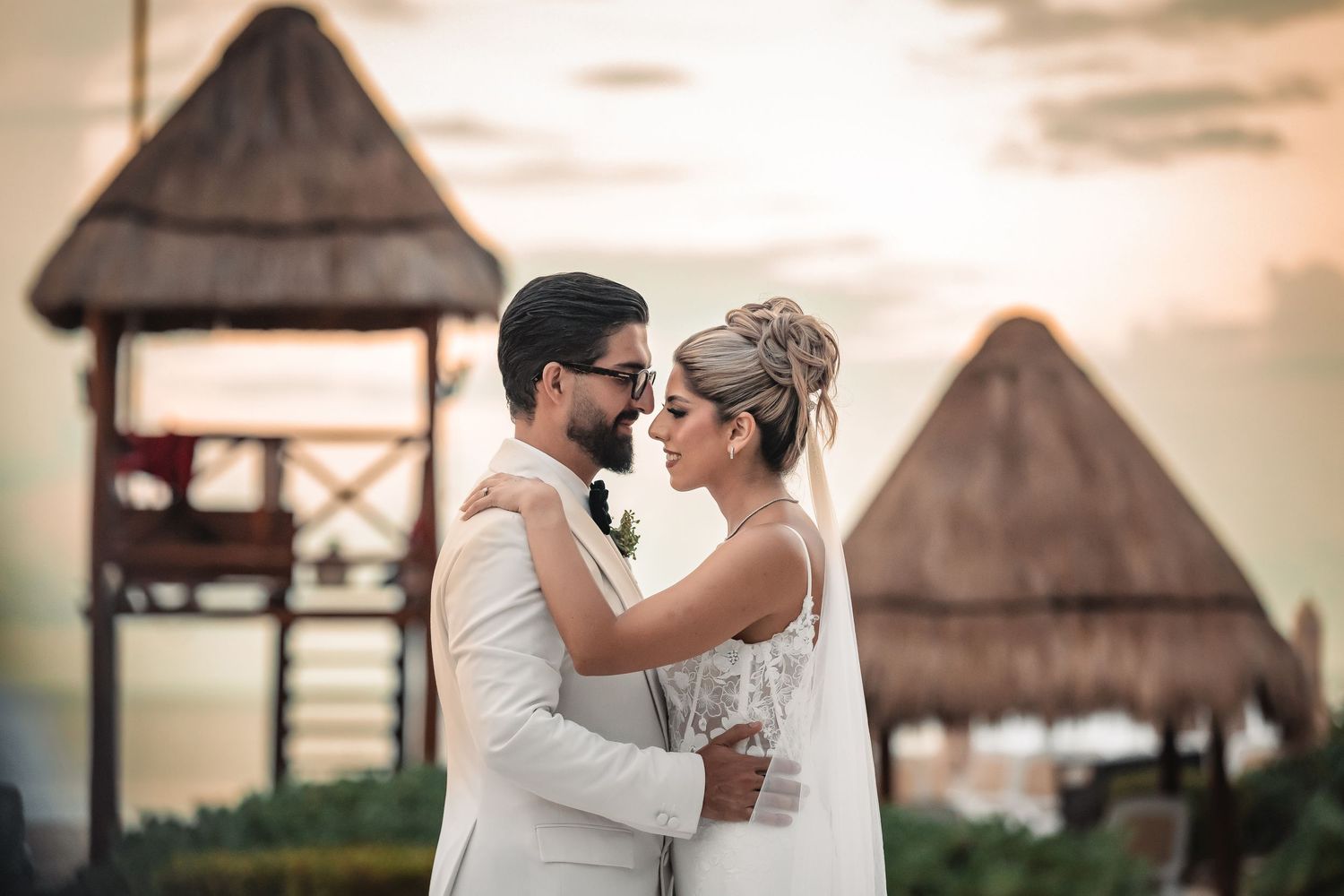 Romantic wedding couple embraces against a sunset sky with thatched roof huts in the background.
